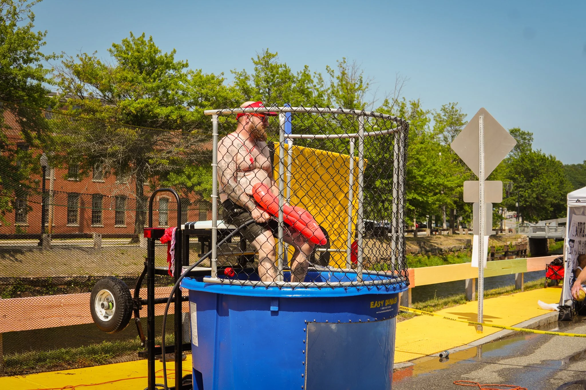 Man in shirt, shorts and red cap standing inside a dunk tank, holding a red dunking seat, outdoors on a sunny day near a water splash on the ground.