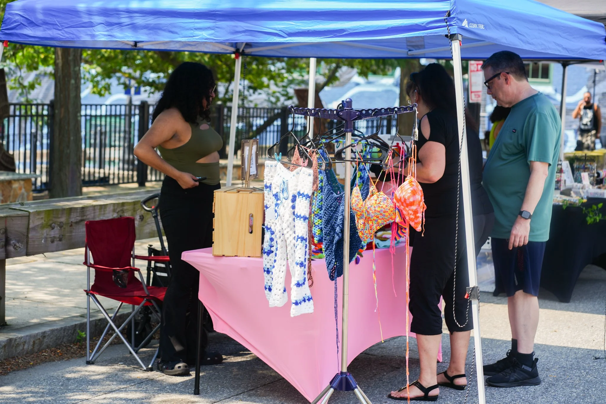 A woman at a craft stall selling colorful crochet items, with two customers browsing, under a blue canopy on a sidewalk.