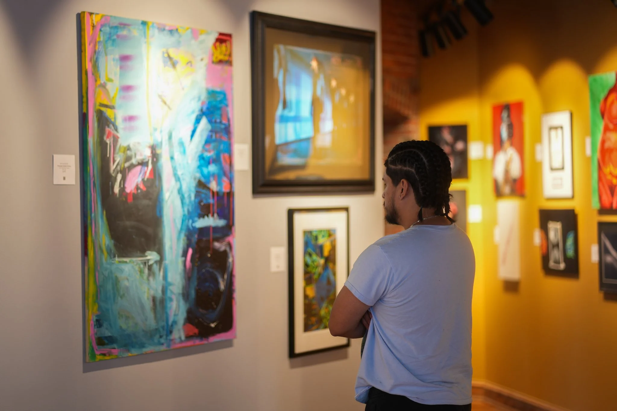 A man with braided hair standing with arms crossed, observing abstract and colorful artwork in an art gallery with a yellow wall.
