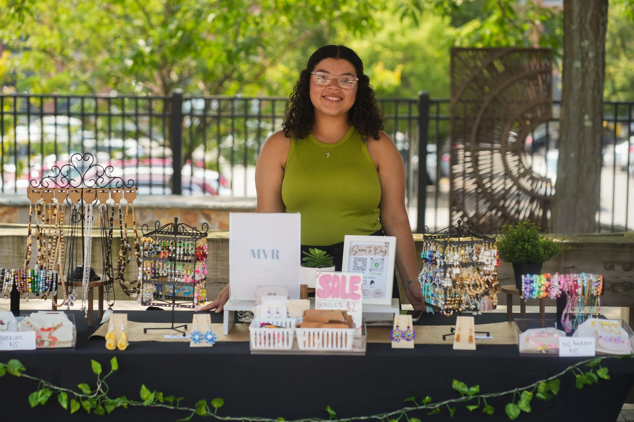 Young woman with glasses smiling at a jewelry stand outdoors, displaying earrings, necklaces, and bracelets for sale.