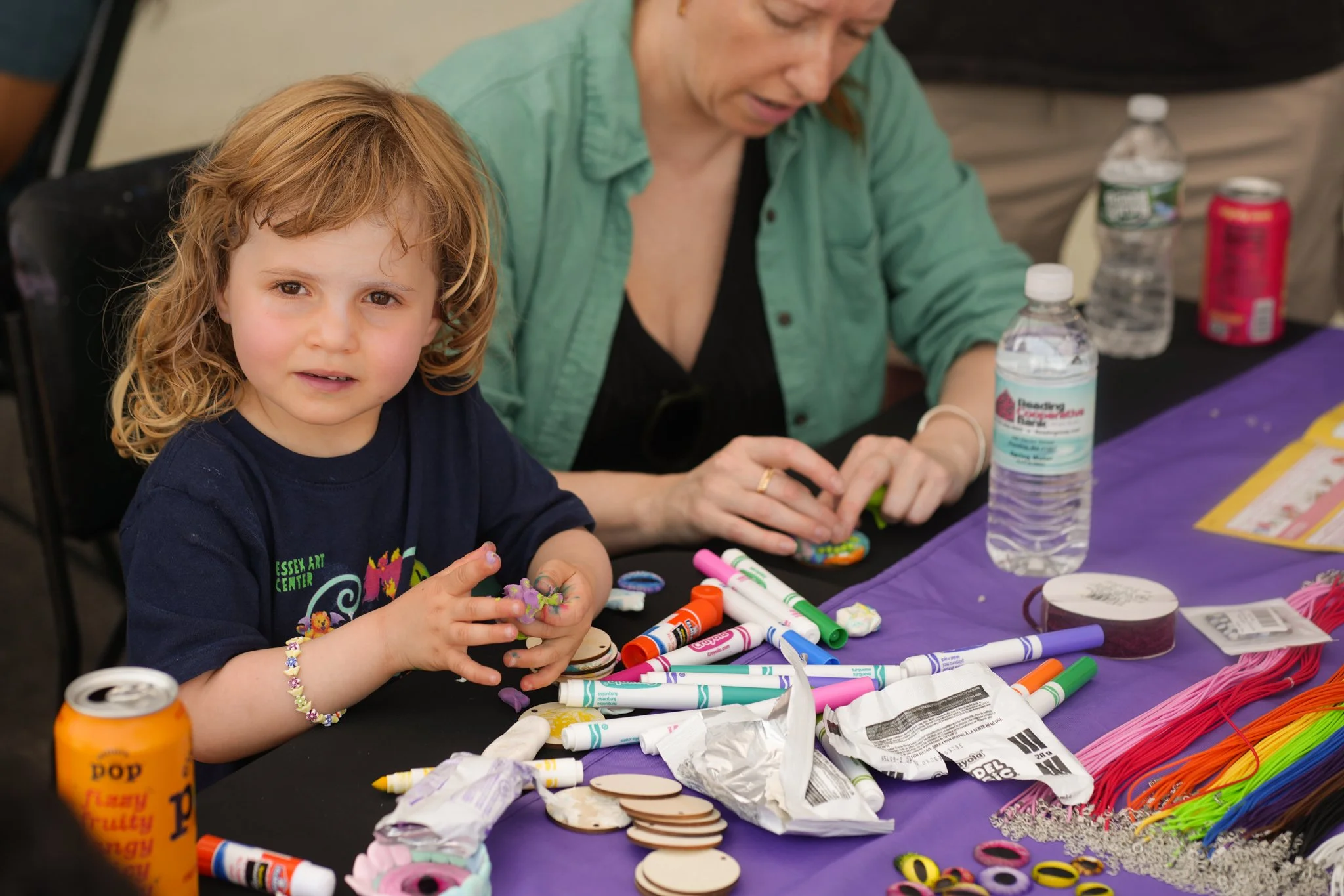 A young girl with curly hair sitting at a table filled with craft supplies, and an adult woman working on a craft project next to her.