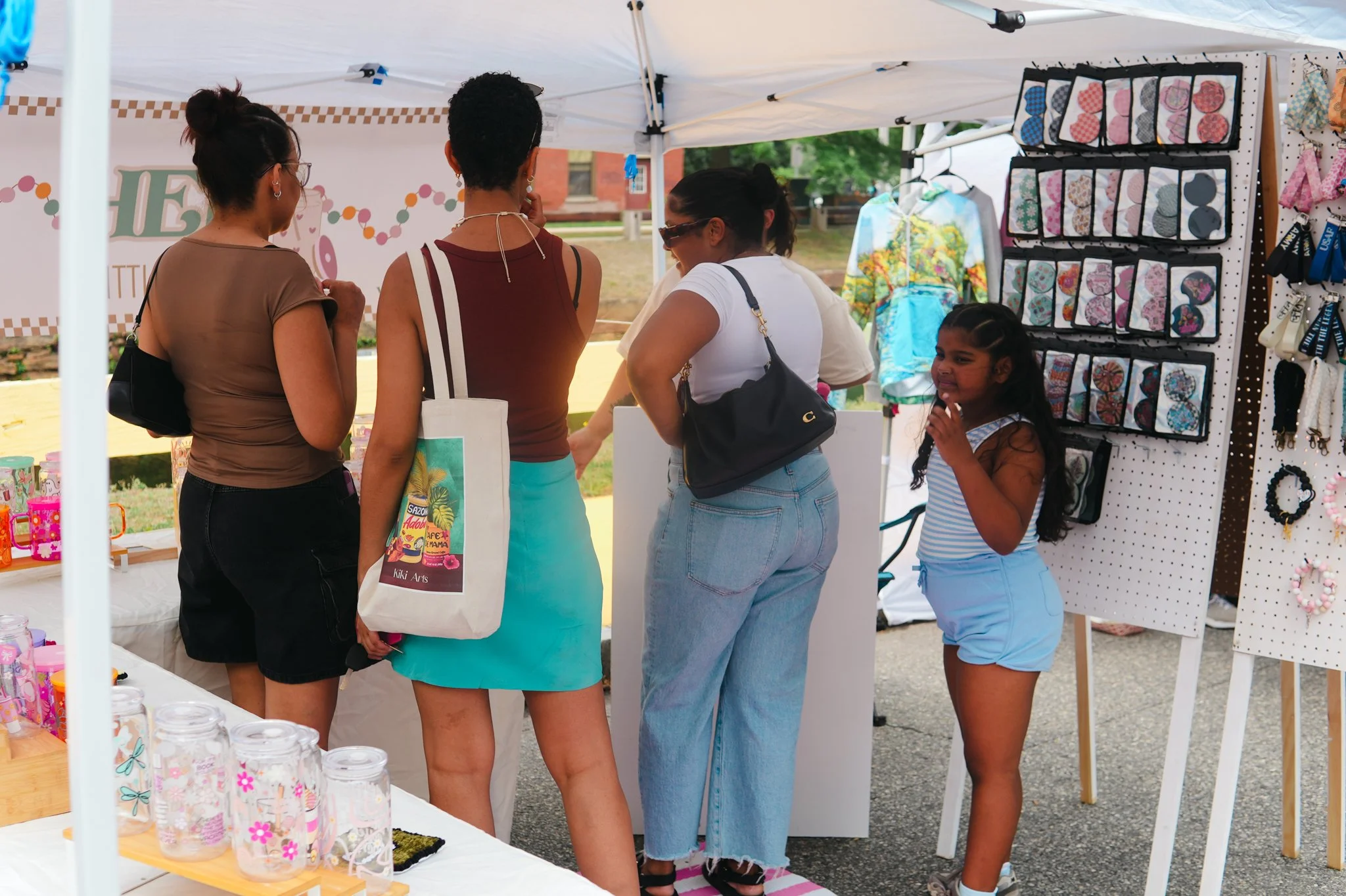 Four women and a young girl looking at items at an outdoor craft market stall with jewelry and decorative jars.