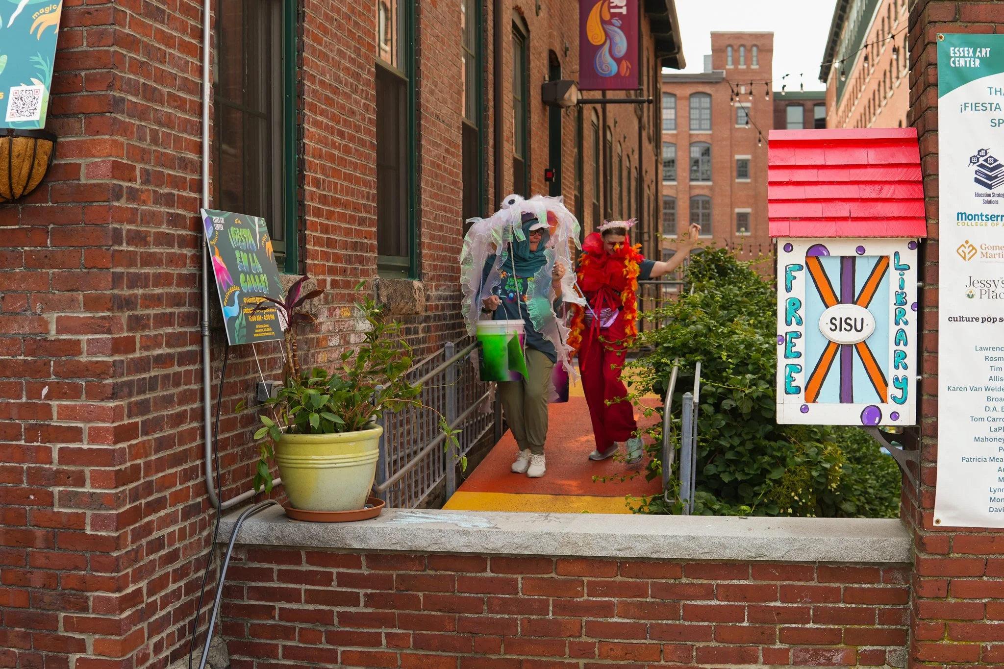 Two people dressed in colorful costumes, walking on a small elevated passageway in front of a brick building during a lively outdoor event. One is wearing a teal hoodie and a whimsical, white, cloud-like headpiece, while the other is dressed in red w