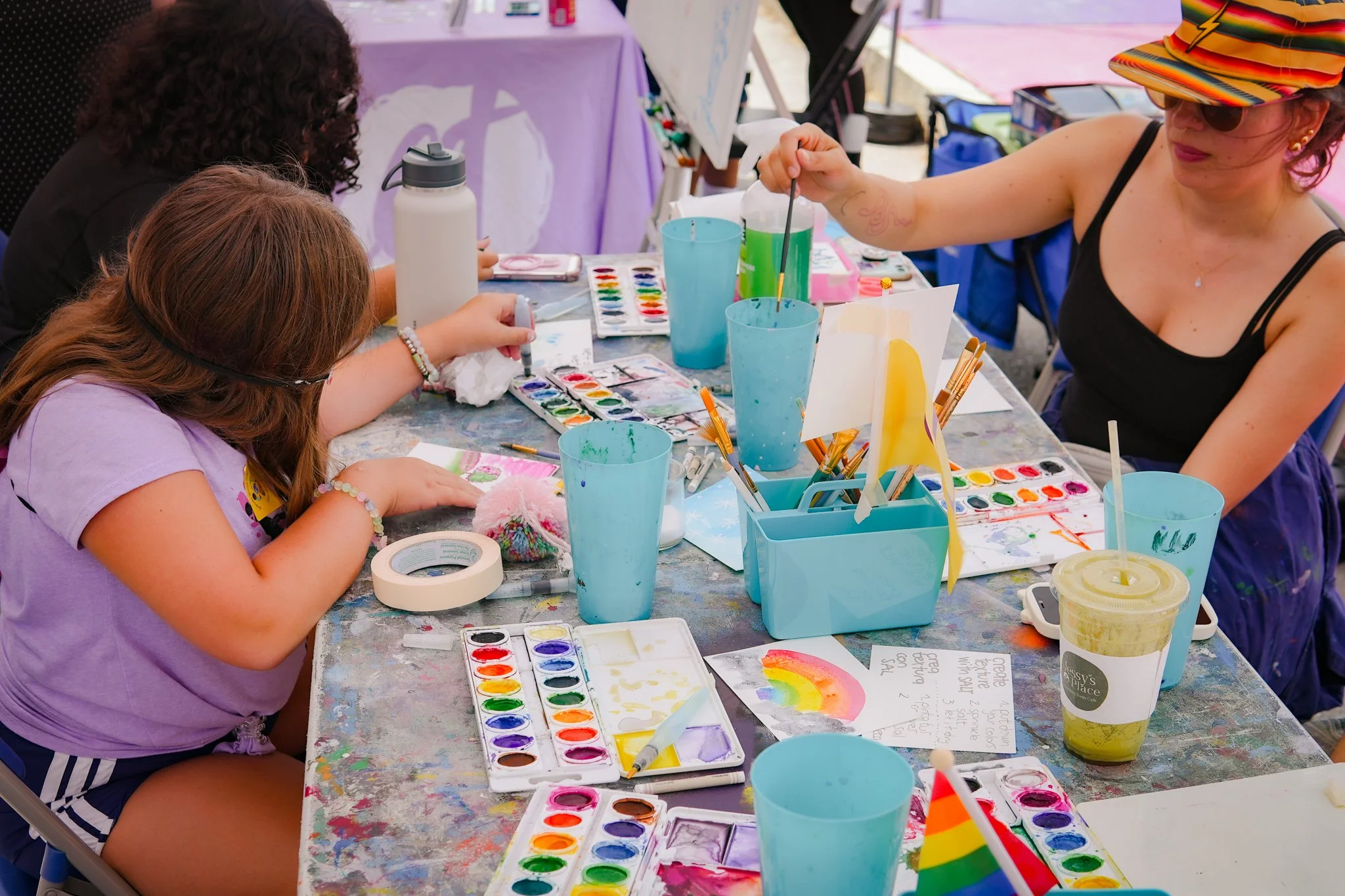 People painting at an outdoor table with watercolors, paintbrushes, and colorful rainbow-themed artwork.