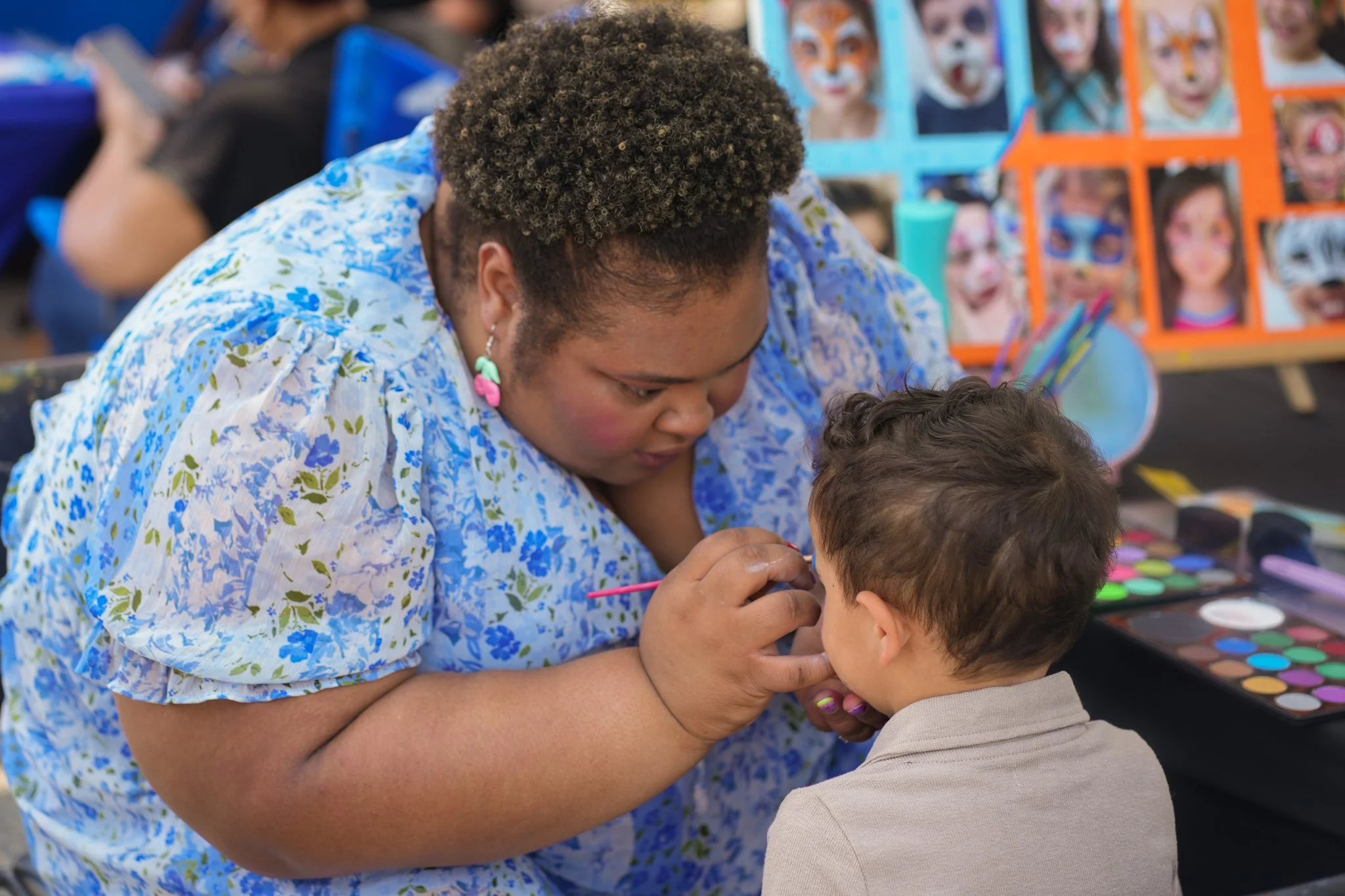 A woman with short curly hair and colorful earrings is face painting a young boy at a fair or carnival, with face painting supplies on the table and colorful face paintings displayed in the background.