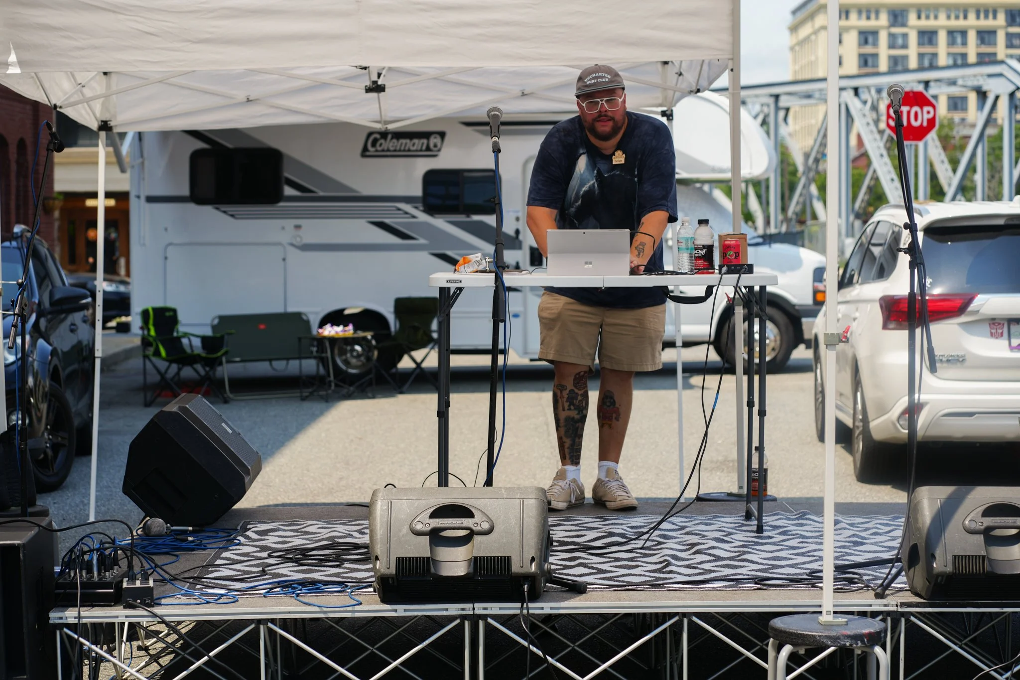 A man standing behind a DJ table with equipment, under a white canopy tent, with parked cars and RV in the background.