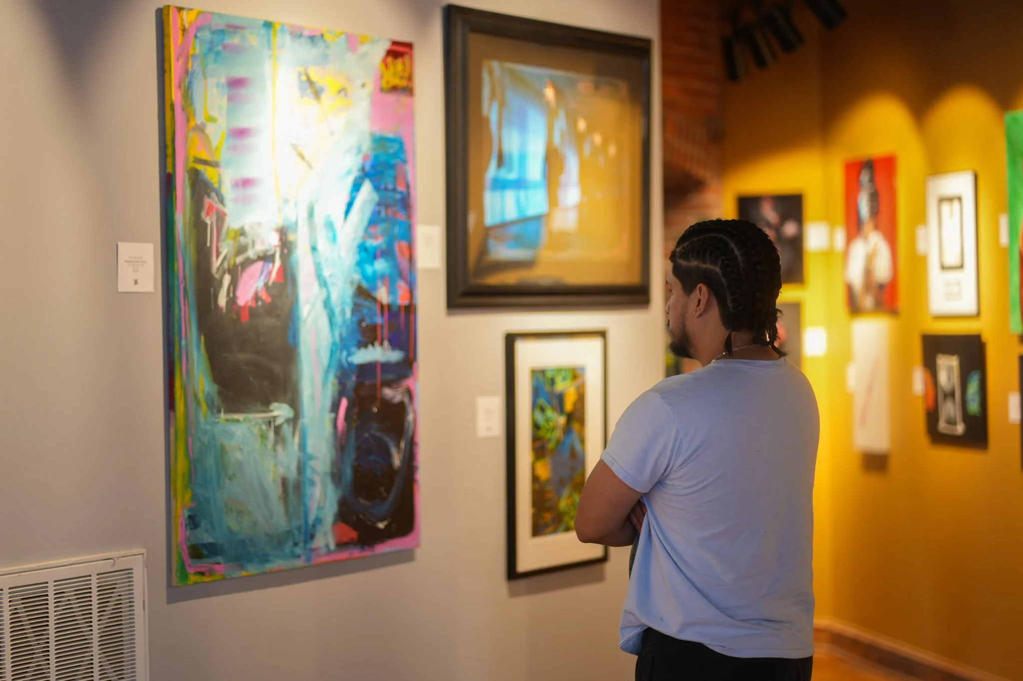 A person with braided hair wearing a light blue T-shirt standing and observing colorful abstract paintings in an art gallery.