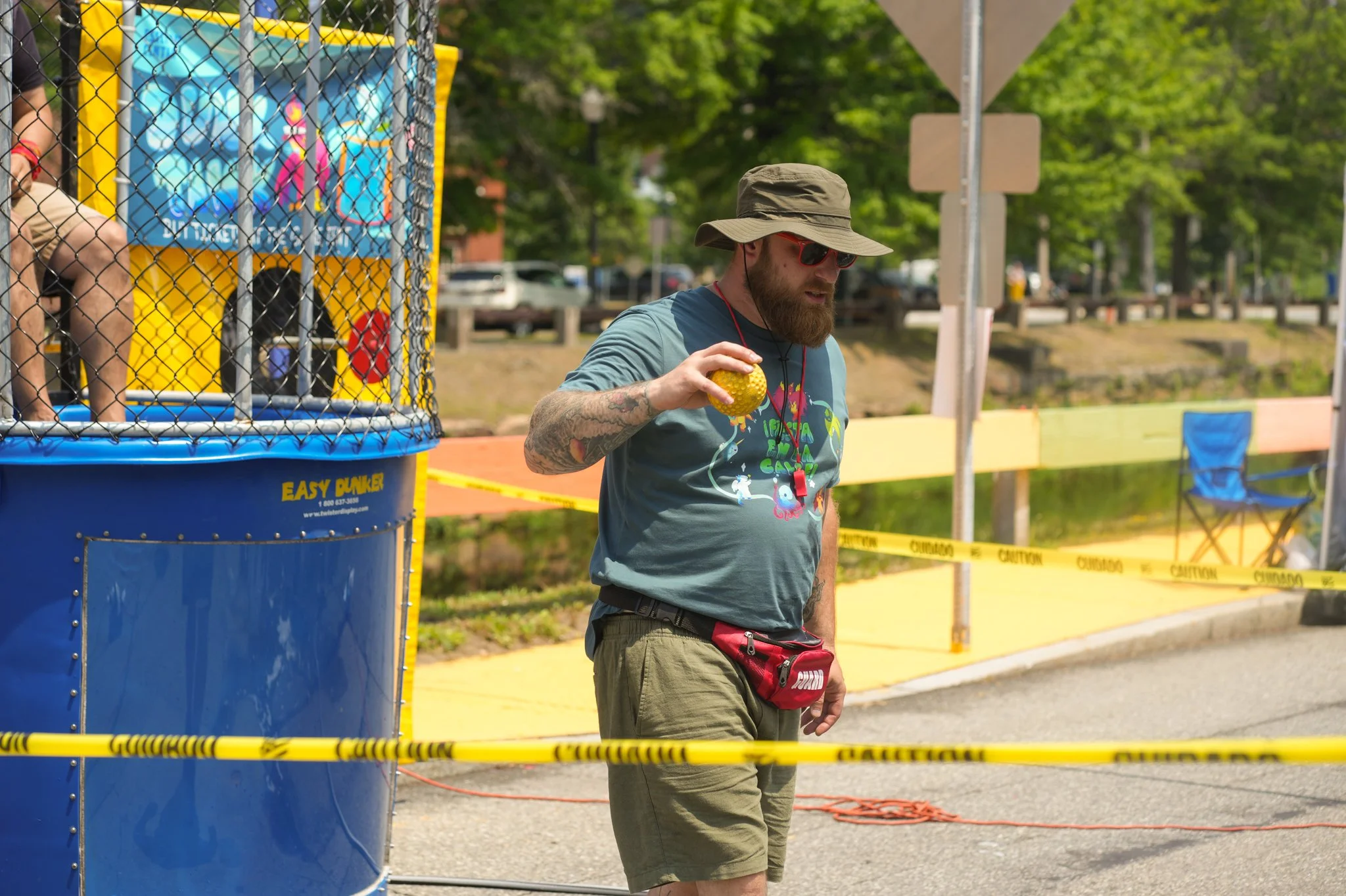 A man with a beard, sunglasses, and a wide-brimmed hat standing outdoors holding a yellow ball, near a blue and yellow dunk tank at a concert or festival site with caution tape, chairs, and trees in the background.