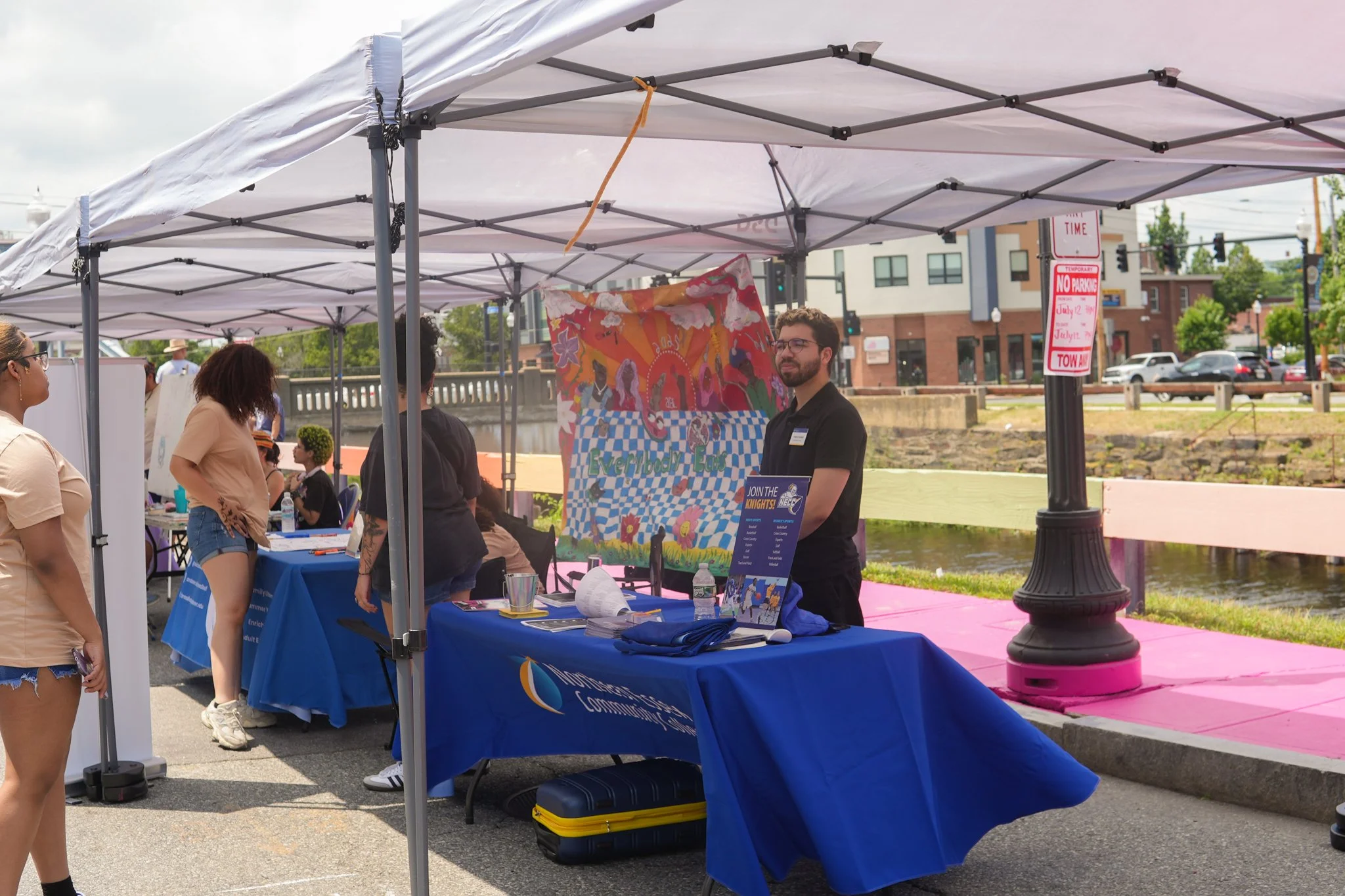 A community event booth with a blue tablecloth, a colorful banner featuring pink flamingos, and people gathered under a white canopy outdoors near a river with buildings in the background.