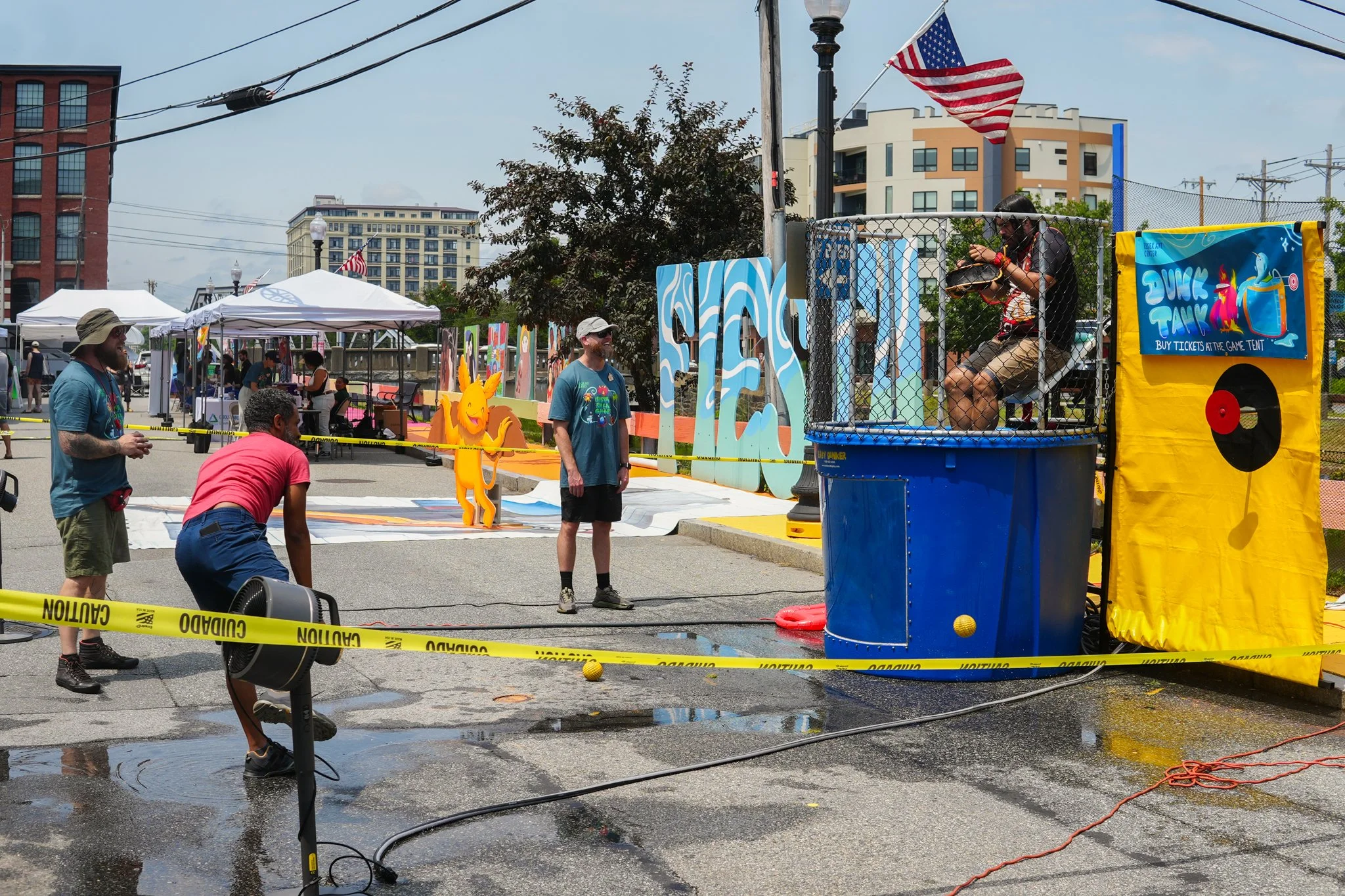 People playing a dunk tank game at an outdoor fair with booths, tents, and colorful decorations in the background.