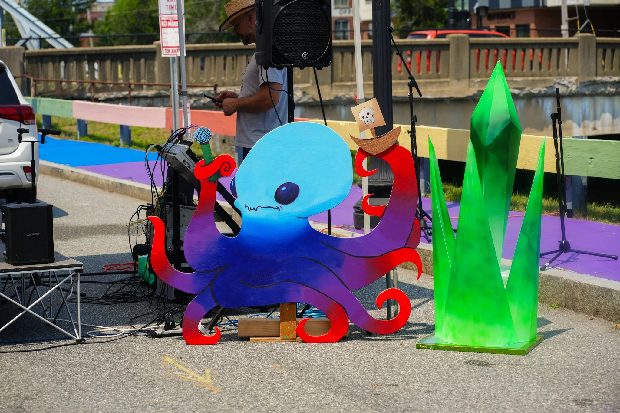 Colorful outdoor display with a large purple octopus and green crystal-shaped sculpture, set on a street with sound equipment, a man in a hat and gray t-shirt, and a bridge in the background.