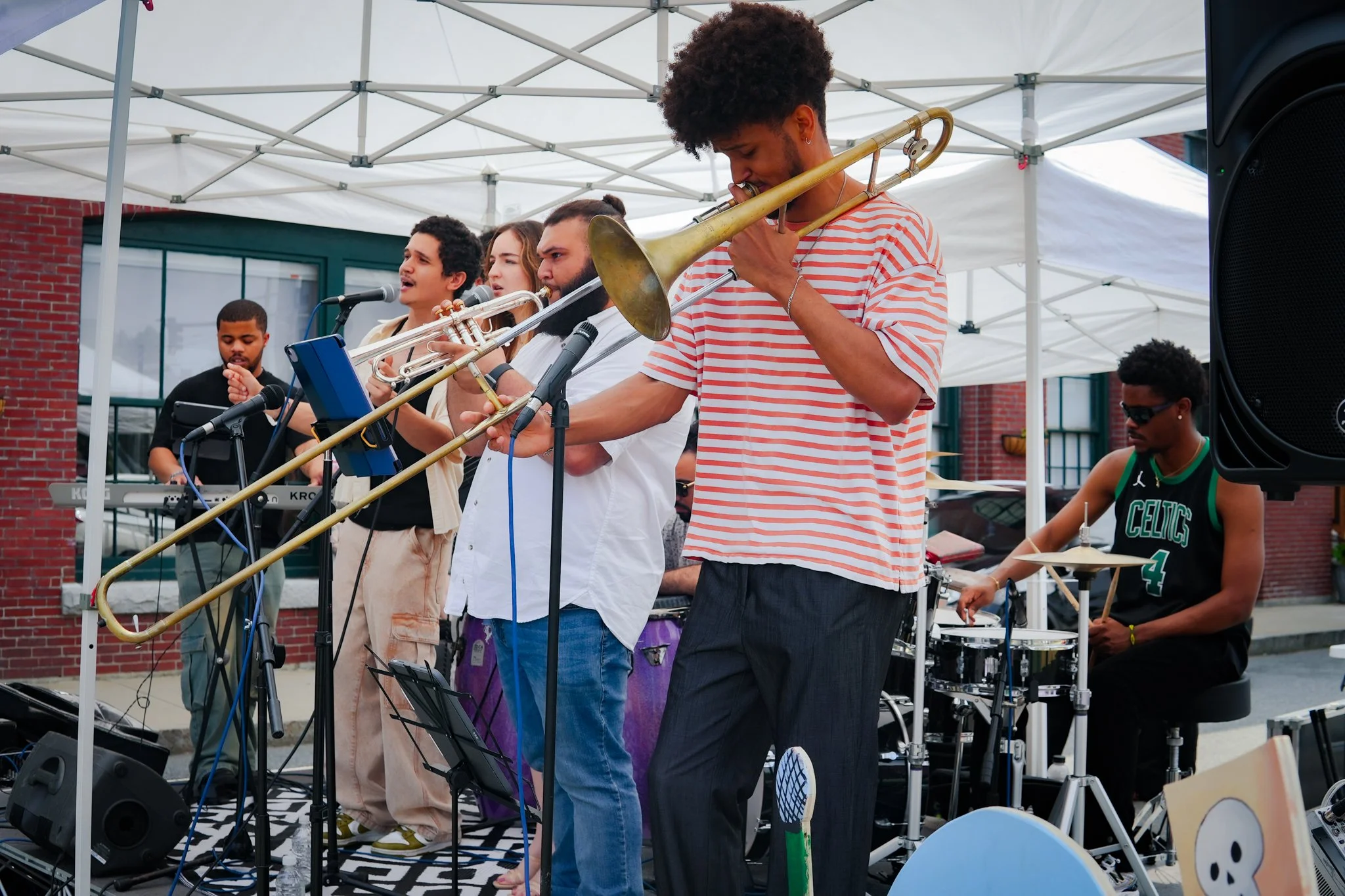 A diverse group of musicians performing outdoors under a white canopy, including a man playing the trumpet, a woman singing into a microphone, and a drummer wearing sunglasses, with various musical equipment around them.