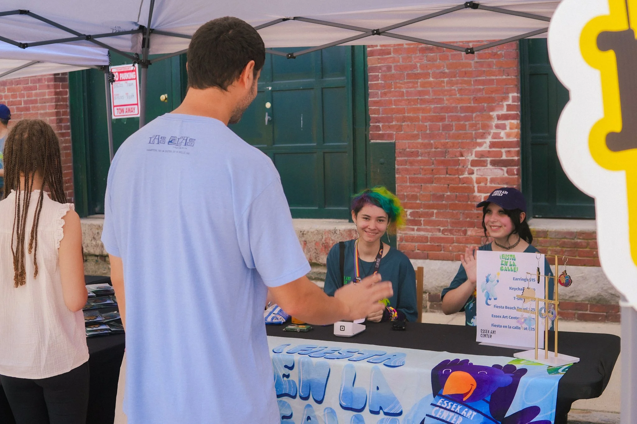 A man is talking to two young women sitting at a booth with a black tablecloth, which displays colorful items and a sign with prices for earrings, keychains, and T-shirts. The women are smiling, one has rainbow-colored hair and is wearing a dark shir