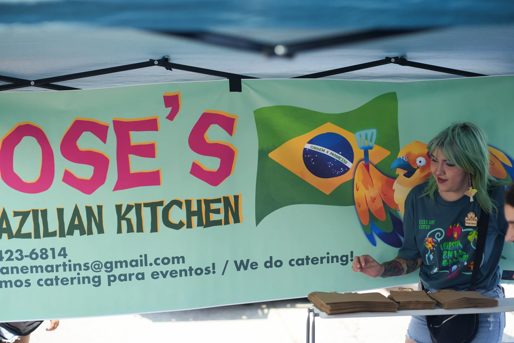 A woman with green hair at a Brazilian-themed food stand with a colorful sign reading 'Ose's Brazilian Kitchen' and a parrot illustration next to the Brazilian flag.