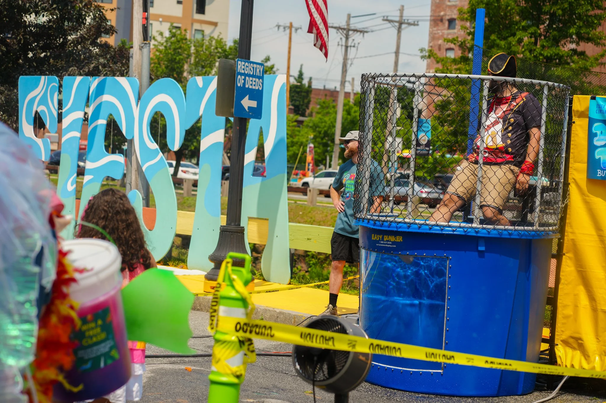 People attending an outdoor event with a water dunk tank and colorful decorations, including large painted blue and green letters and a yellow caution tape in the foreground.