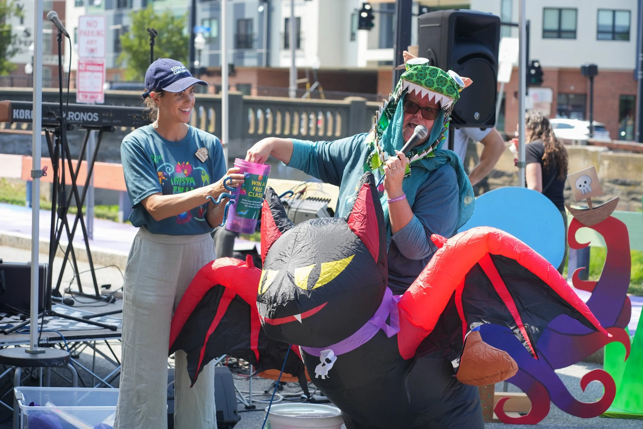Two women at an outdoor event with one dressed in a dragon costume and the other handing her a pink cup. The woman in the costume wears glasses and a hat with dragon features, while the other woman is smiling and wearing a cap and a t-shirt. There is