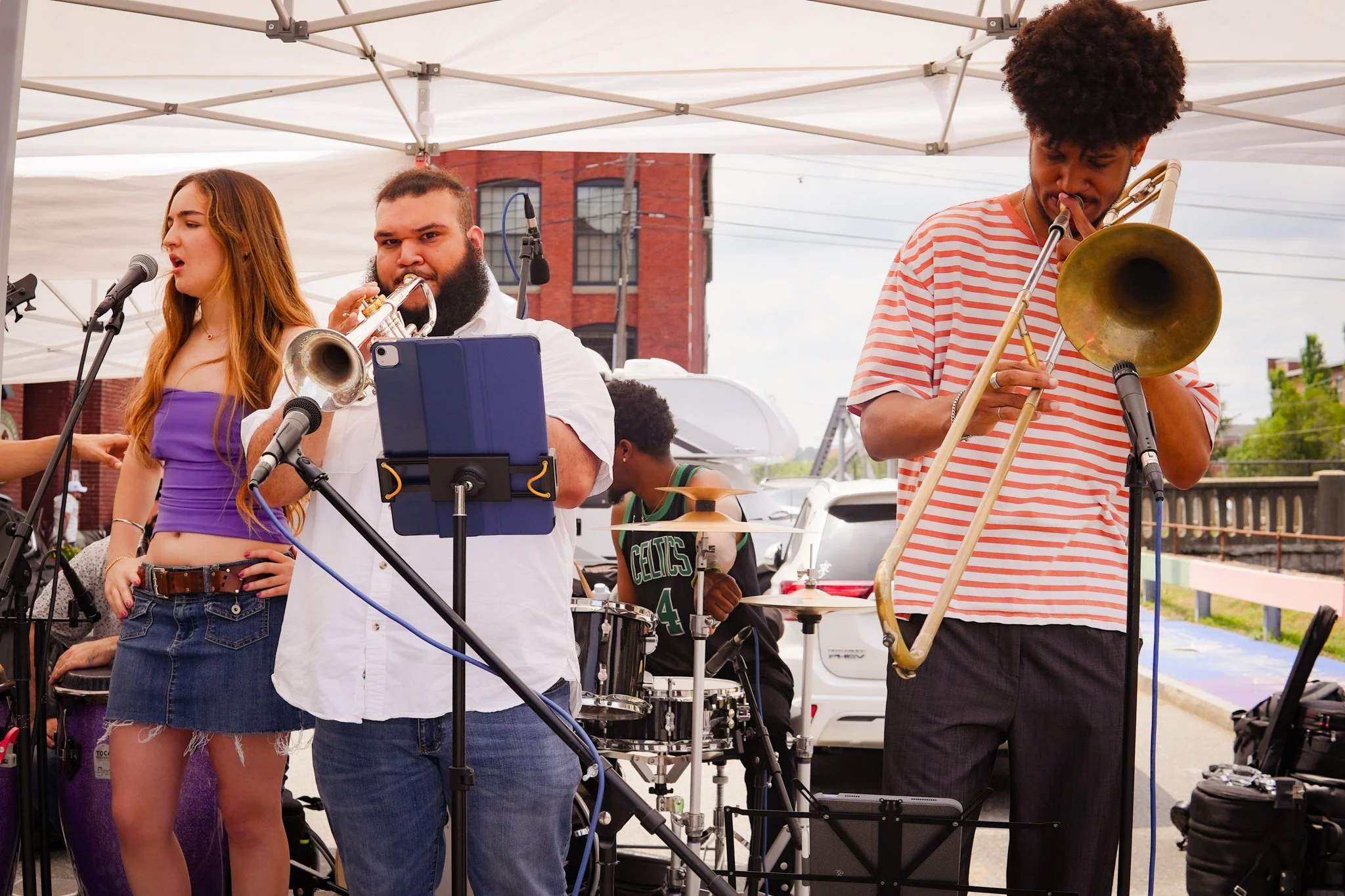 A group of musicians performing outdoors under a canopy, with a woman singing into a microphone, a man playing trumpet, a man with trombone, and a drummer in the background.