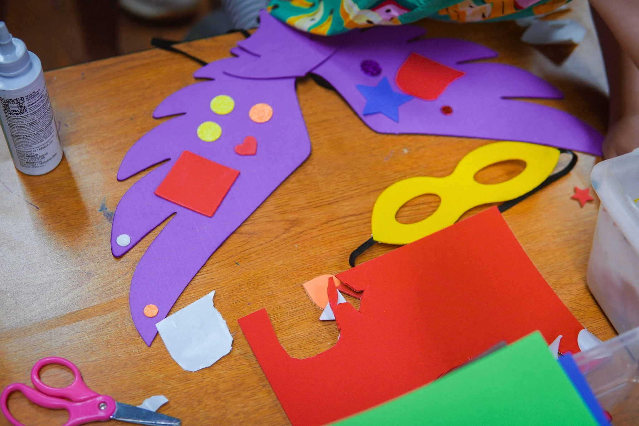 Crafting table with purple paper cutout resembling a crown decorated with colored stickers, a yellow mask, and various pieces of colored paper and scissors.