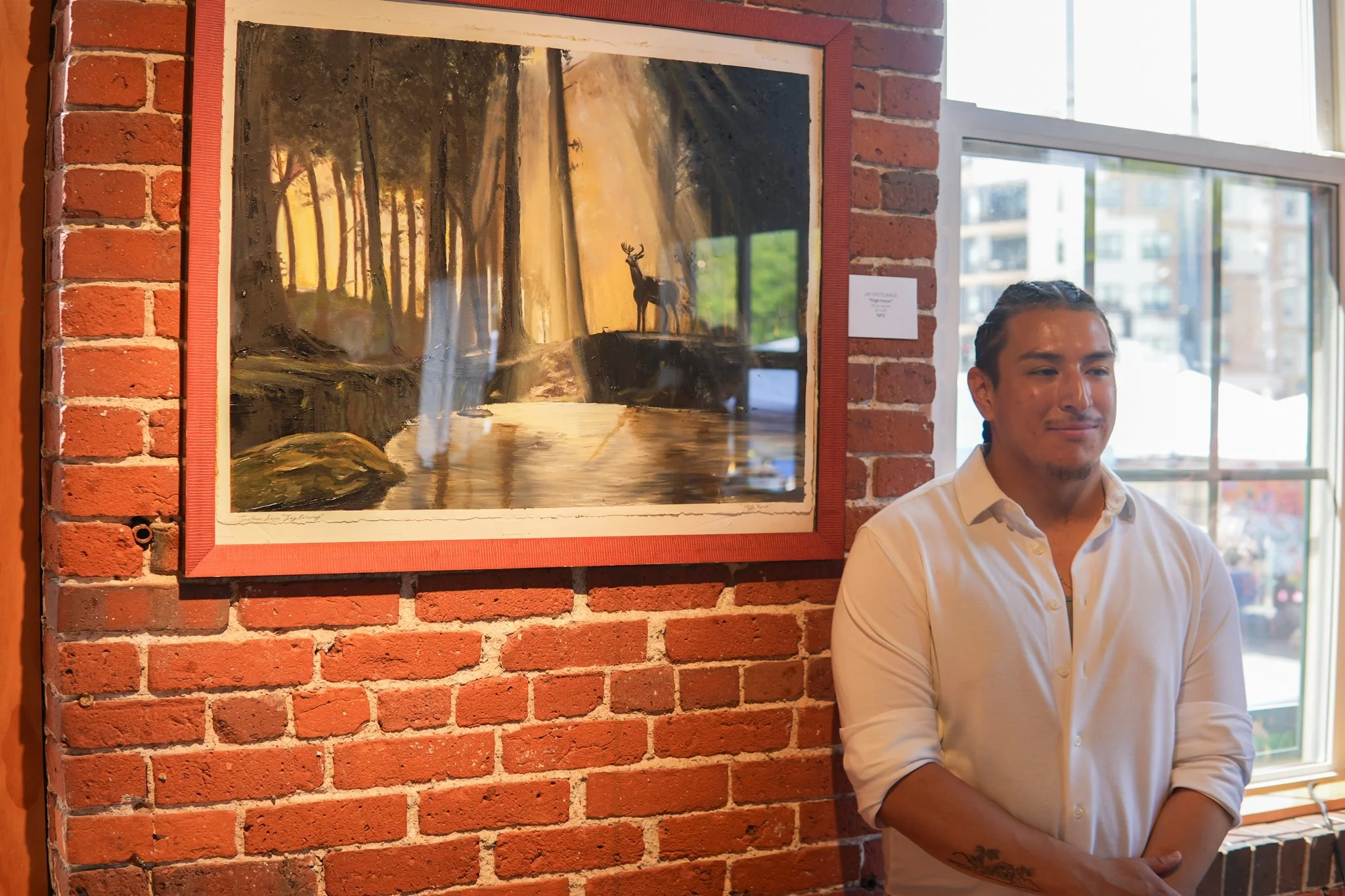 Man standing with arms crossed next to a framed landscape painting of a forest and deer, on a red brick wall inside a gallery with large windows.