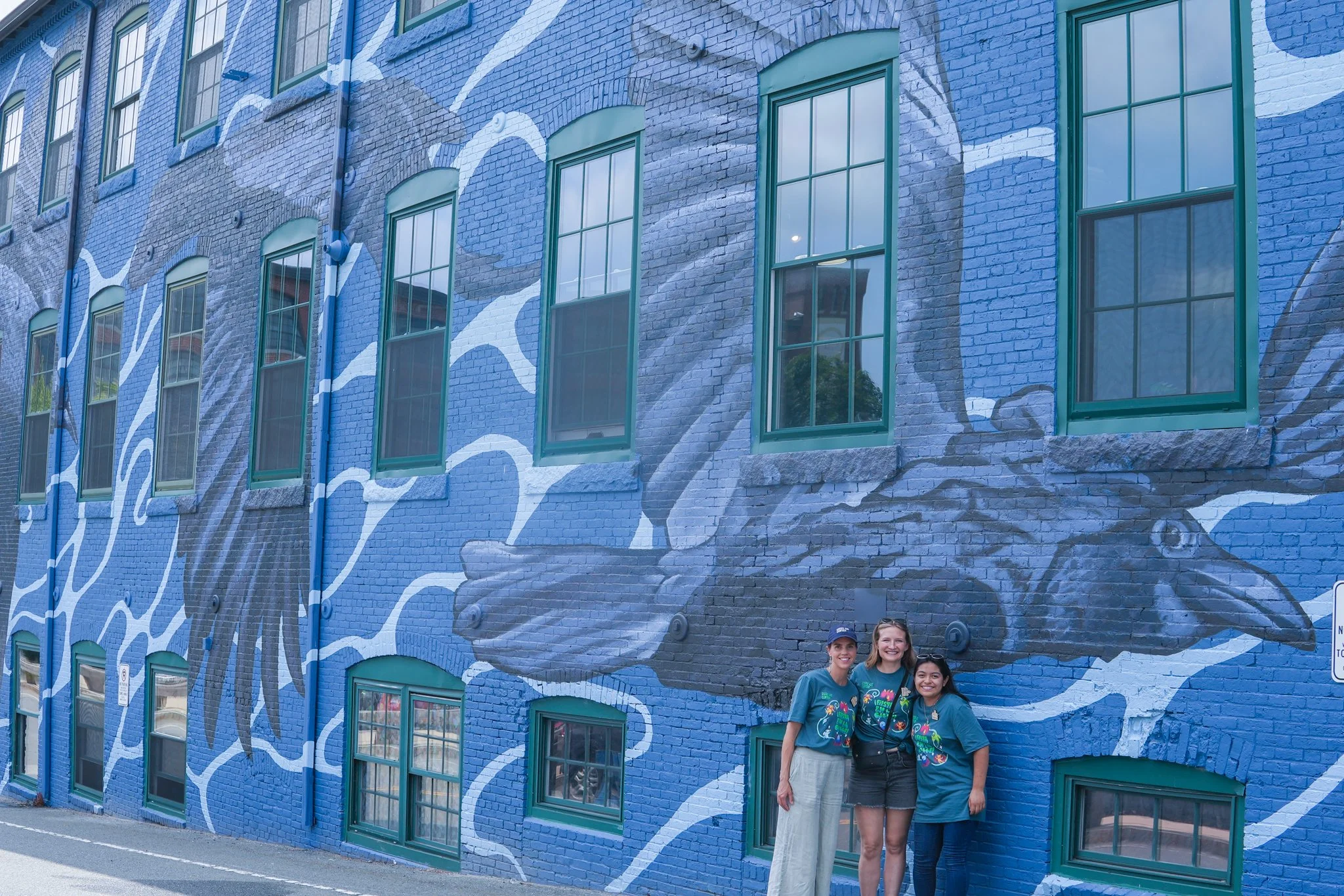 Three women standing in front of a vibrant blue mural of a bird painted on a brick building, with windows incorporated into the artwork.