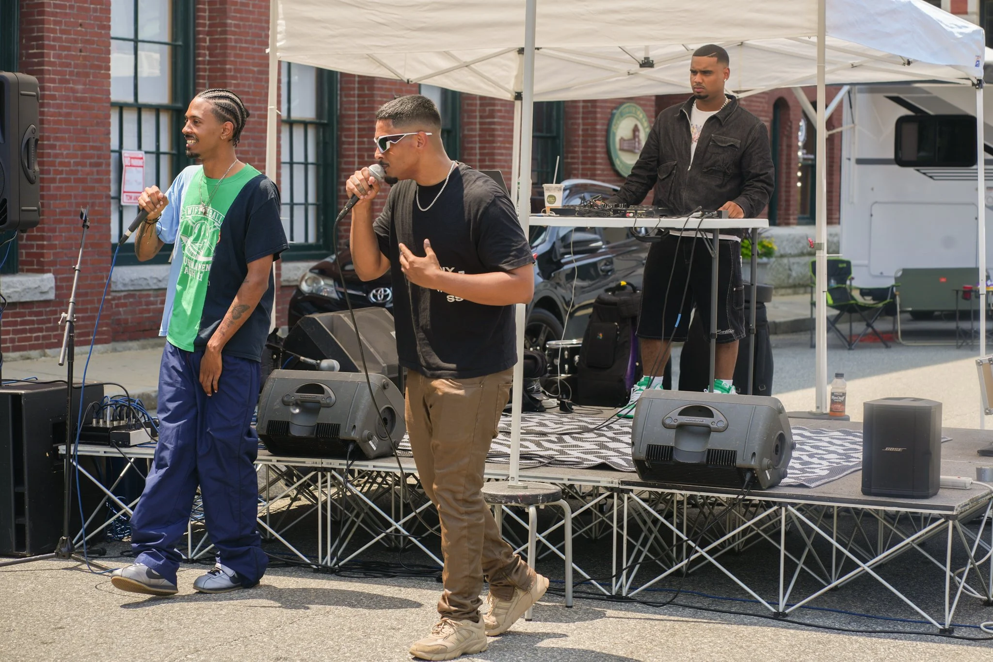 Three men performing music on an outdoor stage inside a city. One man is singing into a microphone, another face shows he is listening, and a third is DJing behind a table with a mixer. The stage is set up in front of red brick buildings.