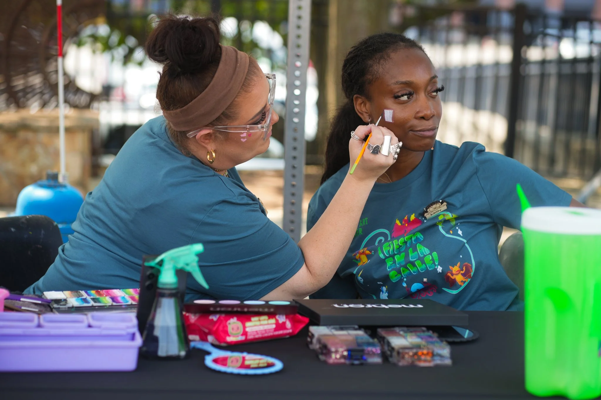 A woman is painting a small purple square on another woman's face during a face painting activity at an outdoor event.