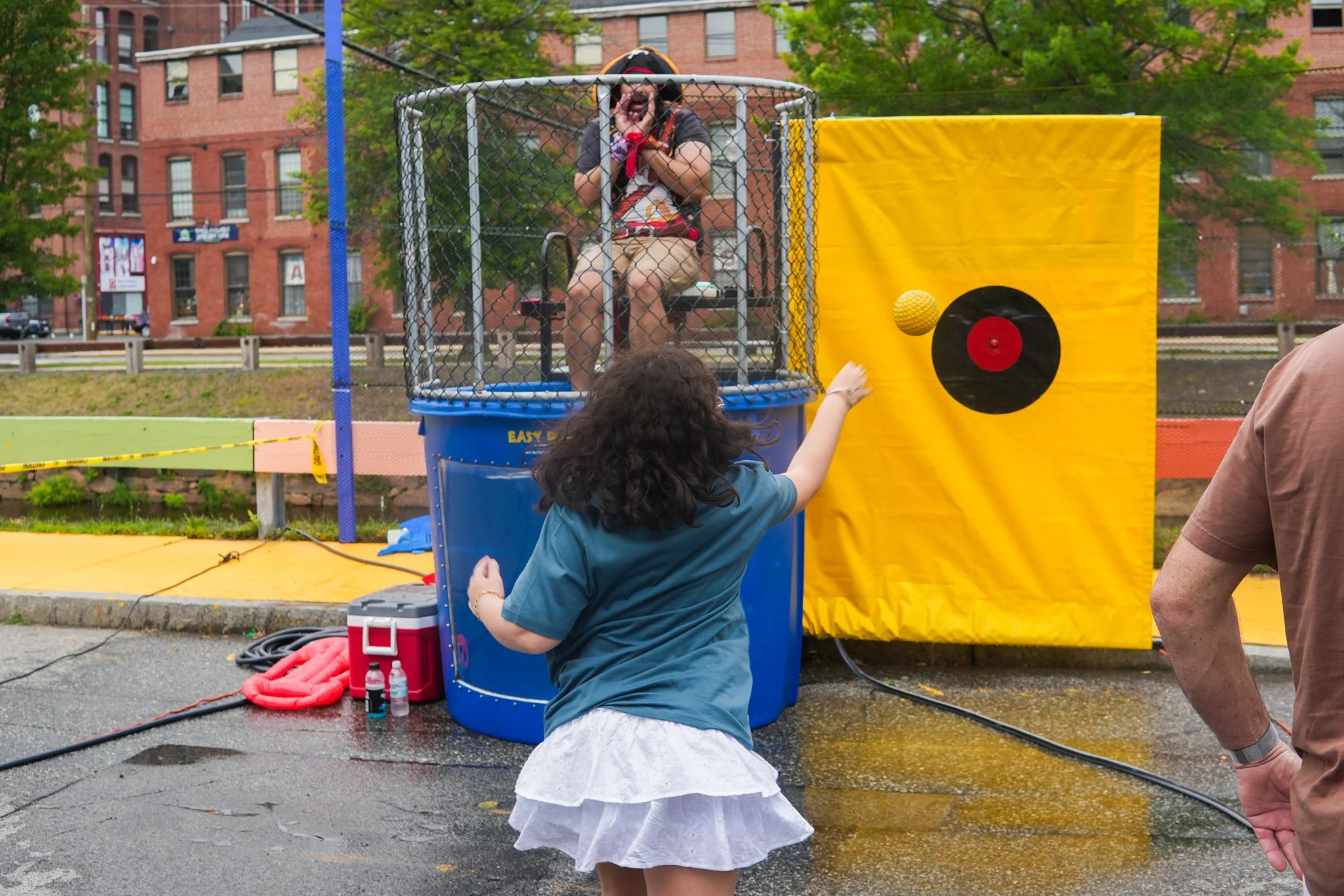 Young girl throwing a ball at a dunk tank with a person inside, during an outdoor event in a city park.