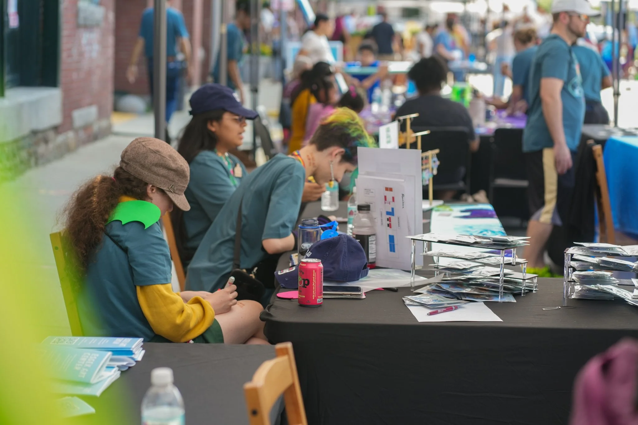 People sitting at tables with displays and pamphlets at an outdoor event or festival.