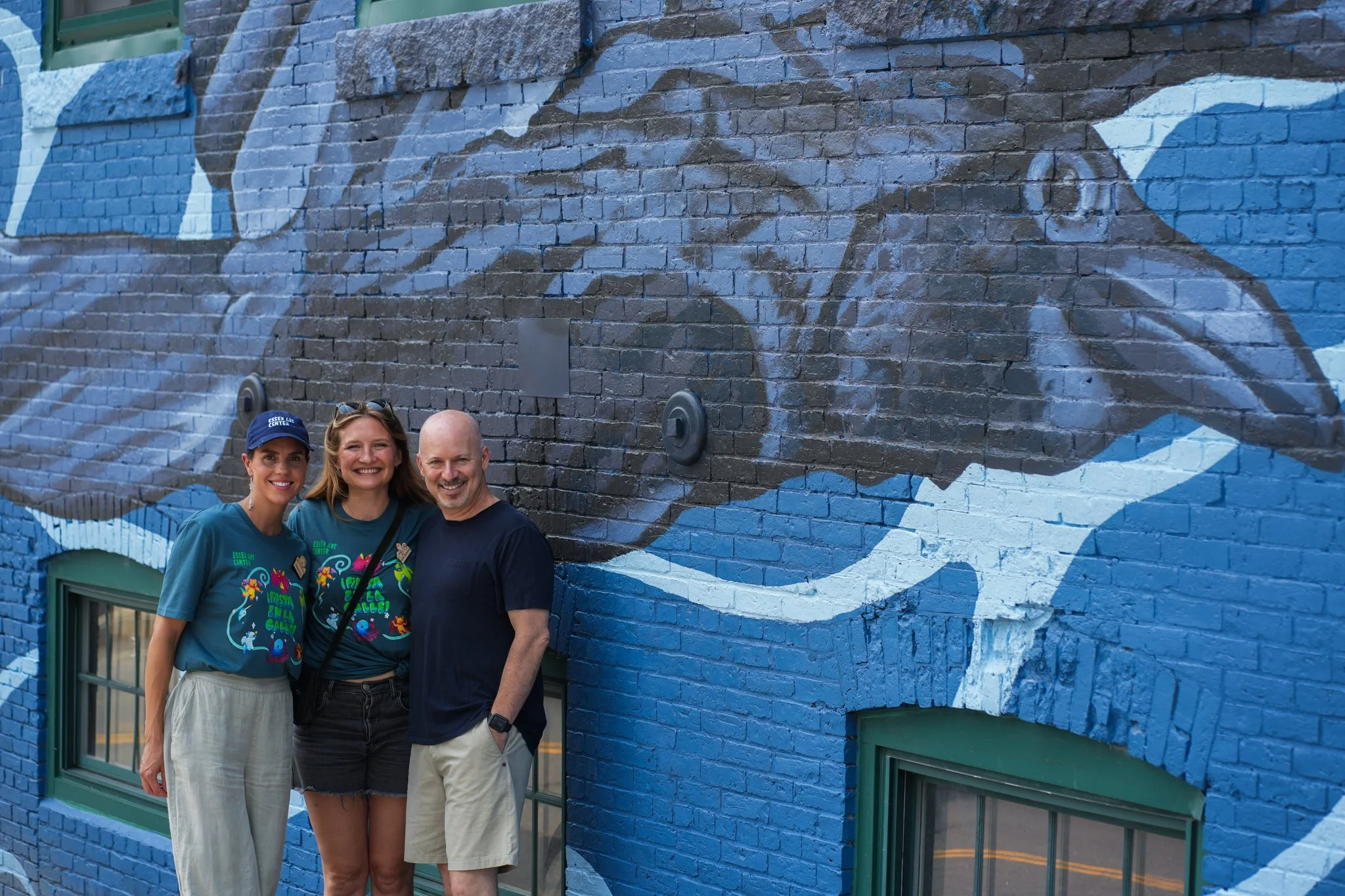 Three people standing in front of a large mural of a toucan painted on a brick wall.