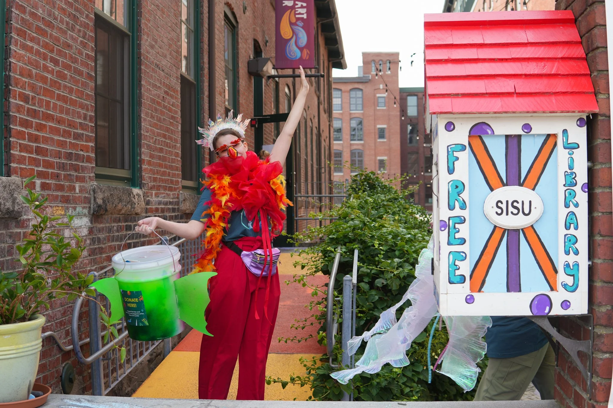 Person dressed in colorful costume with feathers, red scarf, red pants, and glasses, standing outside a brick building next to a donation box labeled 'Tree Library'. The person is holding a green bucket with a label that says 'Donate Here'.
