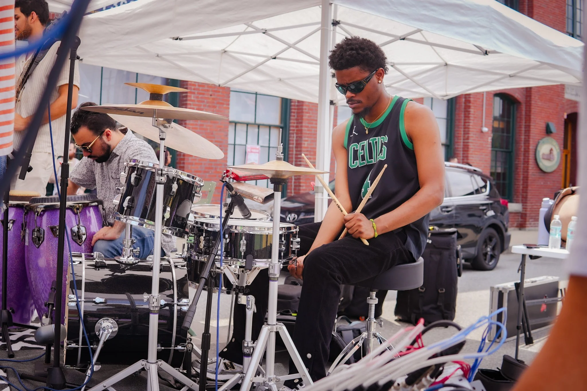 A young man in sunglasses and a black Boston Celtics tank top playing drums at an outdoor event. Other musicians and equipment are visible around him under a white canopy, with a brick building and cars in the background.