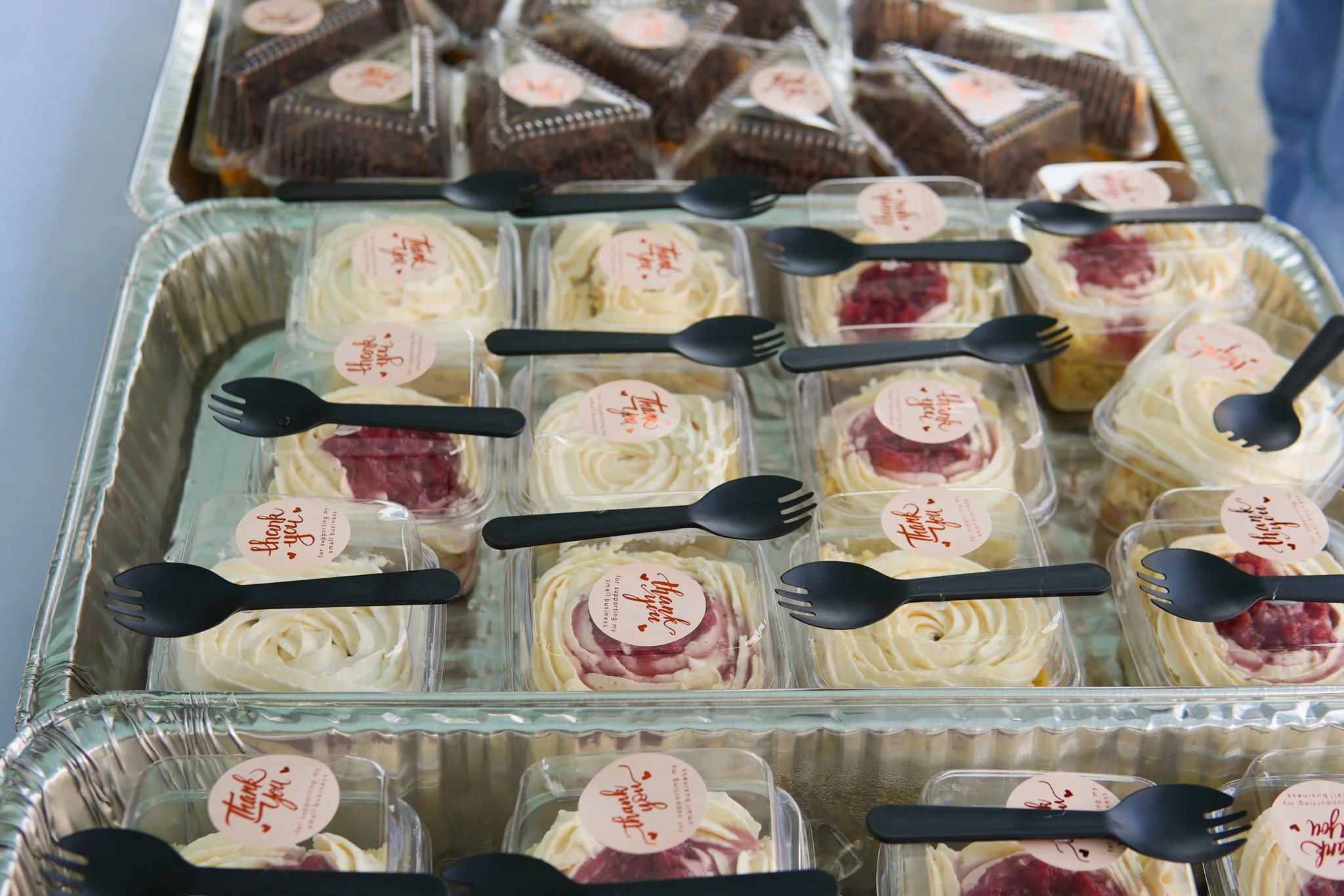 Individual dessert cups with whipped cream and fruit topping, labeled with 'Thank You' stickers, and black plastic forks on a large aluminum tray.