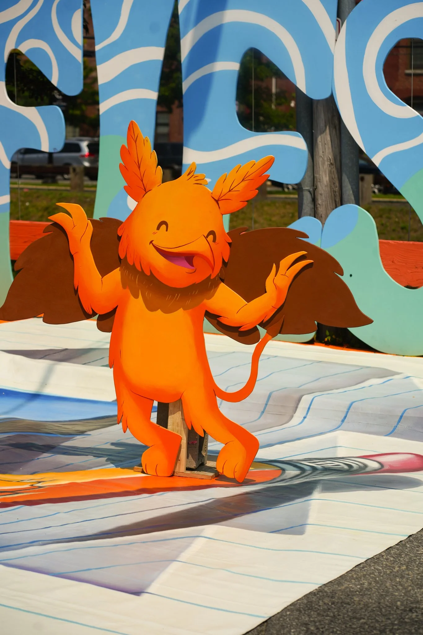 Colorful cartoon lion character with orange fur, large expressive eyes, and a smiling face, standing outdoors with blue decorative background elements and a white surface.