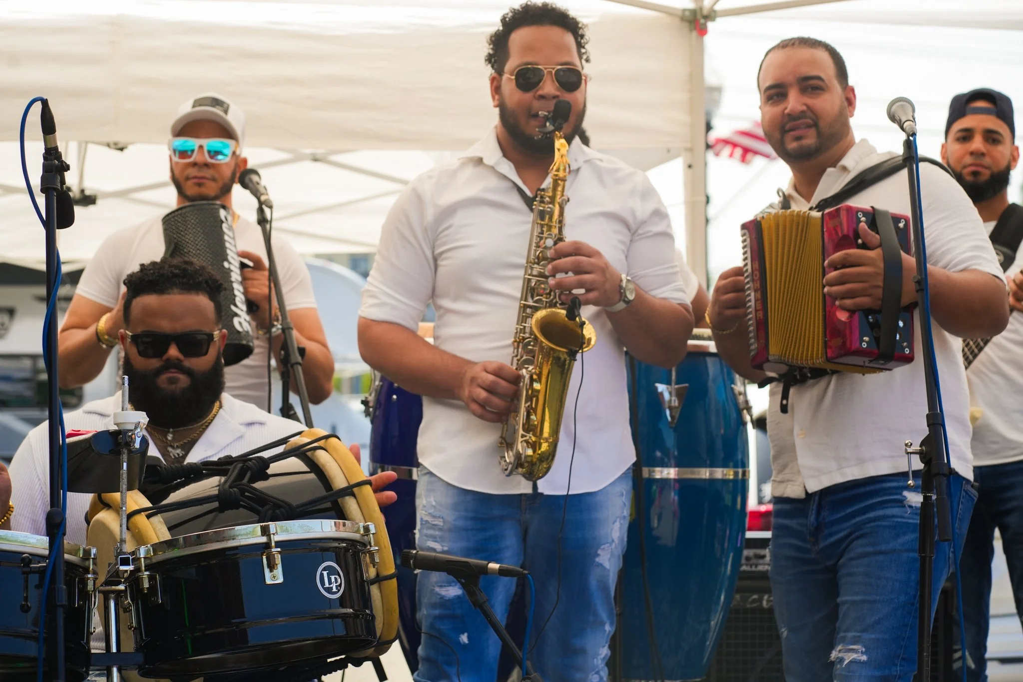 Group of musicians playing instruments outdoors under a white canopy, including a saxophone, accordion, drums, and percussion.