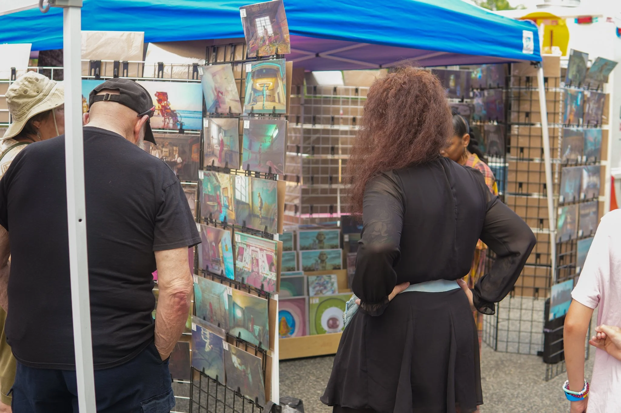 People browsing artwork at an outdoor art market stall.