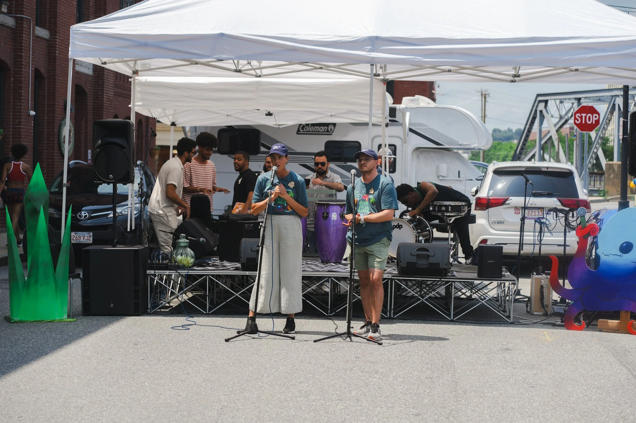 Two people with microphones standing in front of a stage at an outdoor event, with a band setting up and equipment behind them under a white tent.