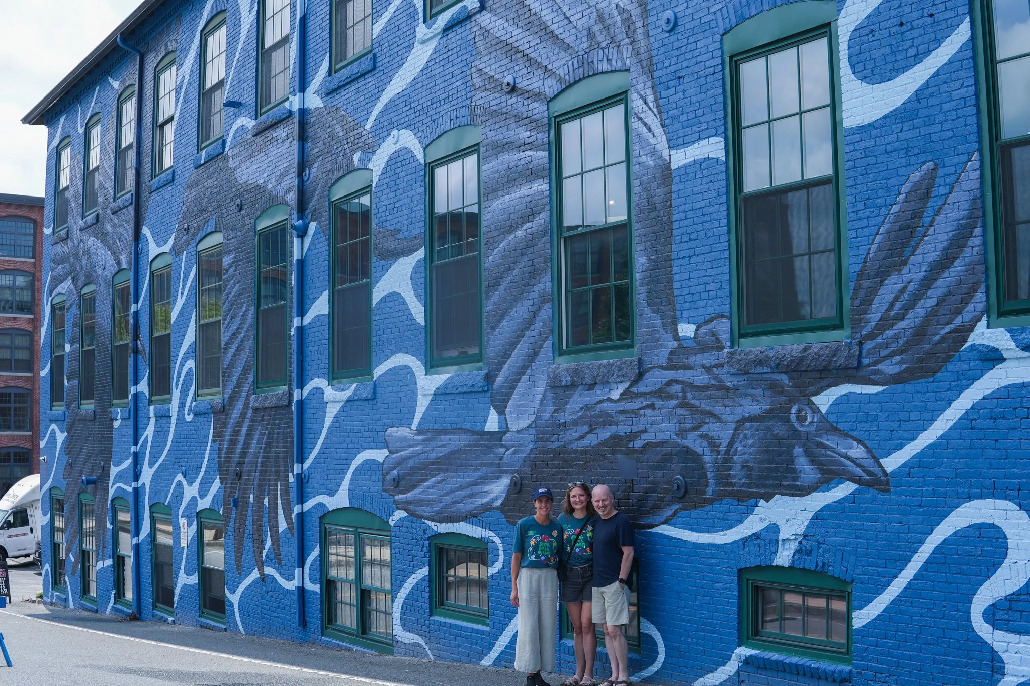 Three people standing in front of a colorful mural on a brick building, featuring a large, detailed depiction of a fish with flowing fins and flowing water details, in shades of blue and gray.