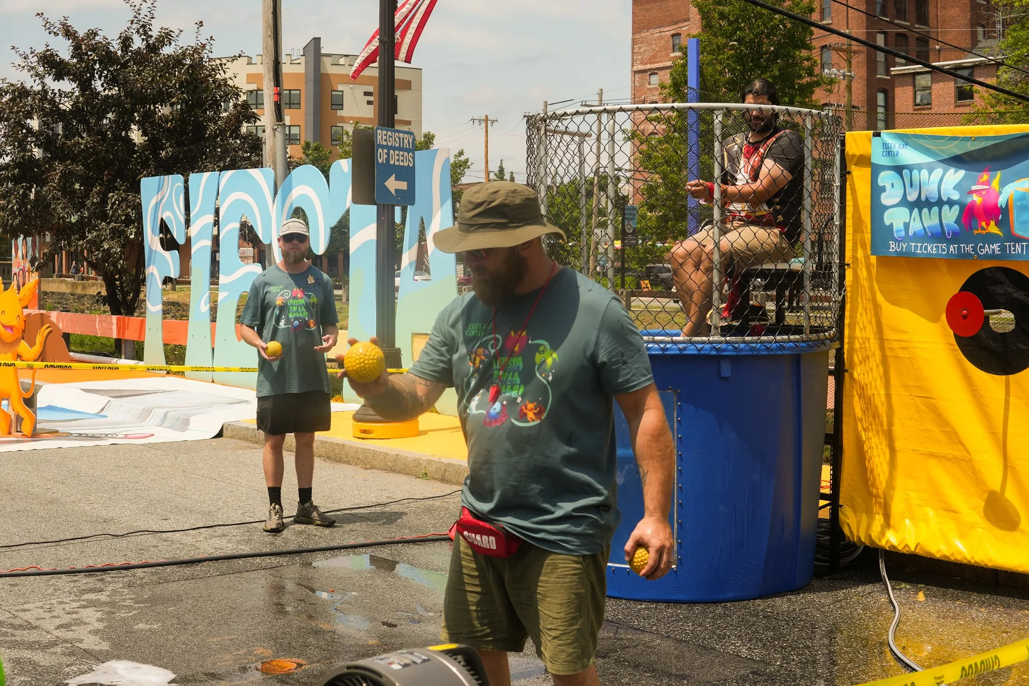 Two men participate in a game at a street carnival on a sunny day. One man is in front, balancing three yellow balls, while the other is standing in the background holding a yellow ball. A woman is seated inside a small cage platform, overseeing the
