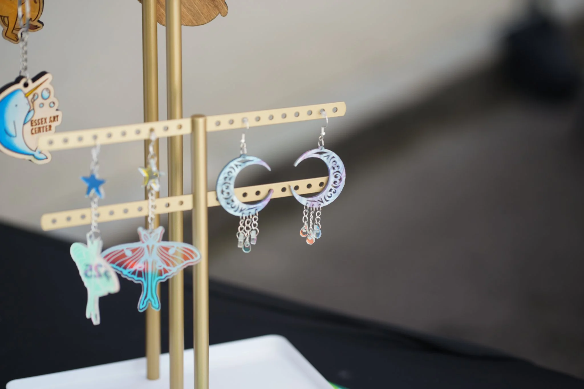 Earrings and charms hanging from a display stand at an art fair, with a sign reading 'Essex Art Center' in the background.