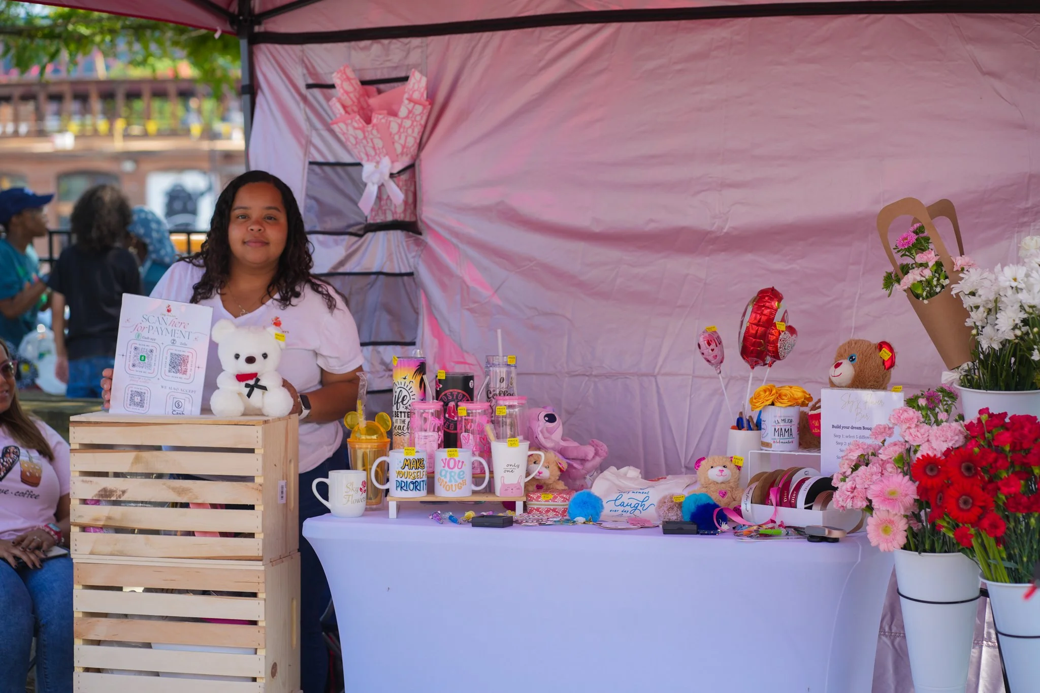 Girl standing behind a table of colorful gifts and flowers at a market stall.