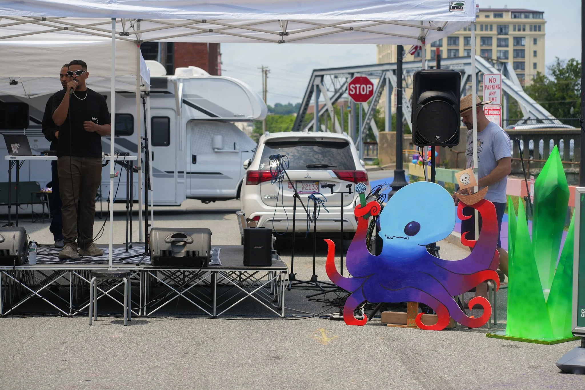 A man with sunglasses is on a small outdoor stage, speaking or singing into a microphone, with a DJ behind him. There are colorful cartoon-like decorations of an octopus and tall green grass nearby. In the background, RVs, a white car, and a bridge a
