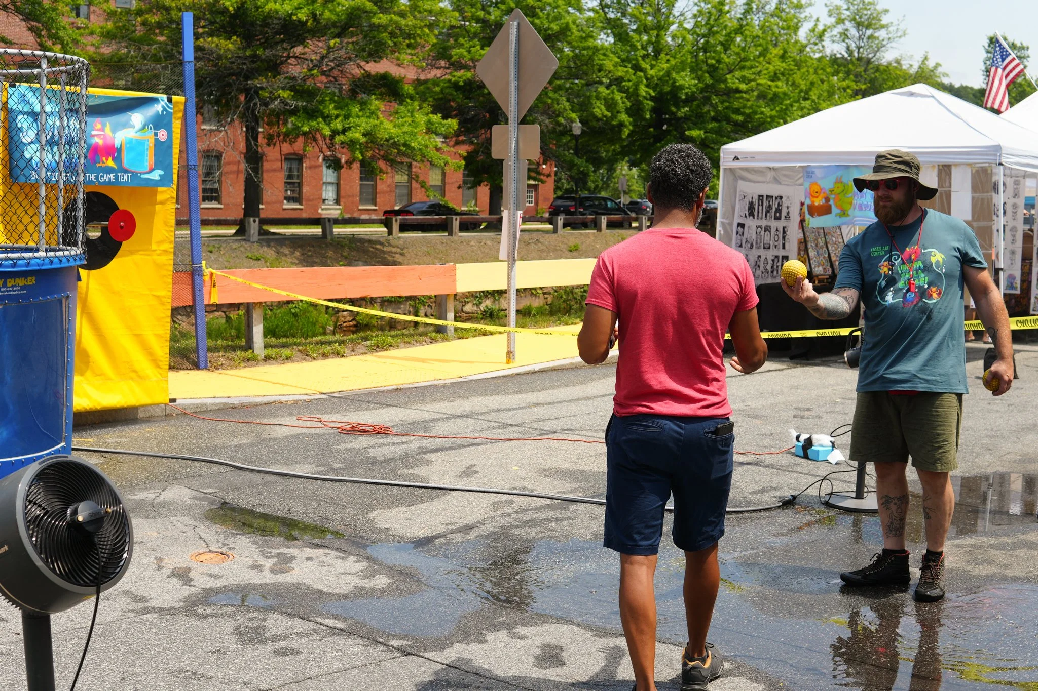 Two men are talking at an outdoor fair or festival, one holding a yellow ball and the other with his back to the camera. There are tents, a game booth, and caution tape in the background, along with a wet pavement and trees.