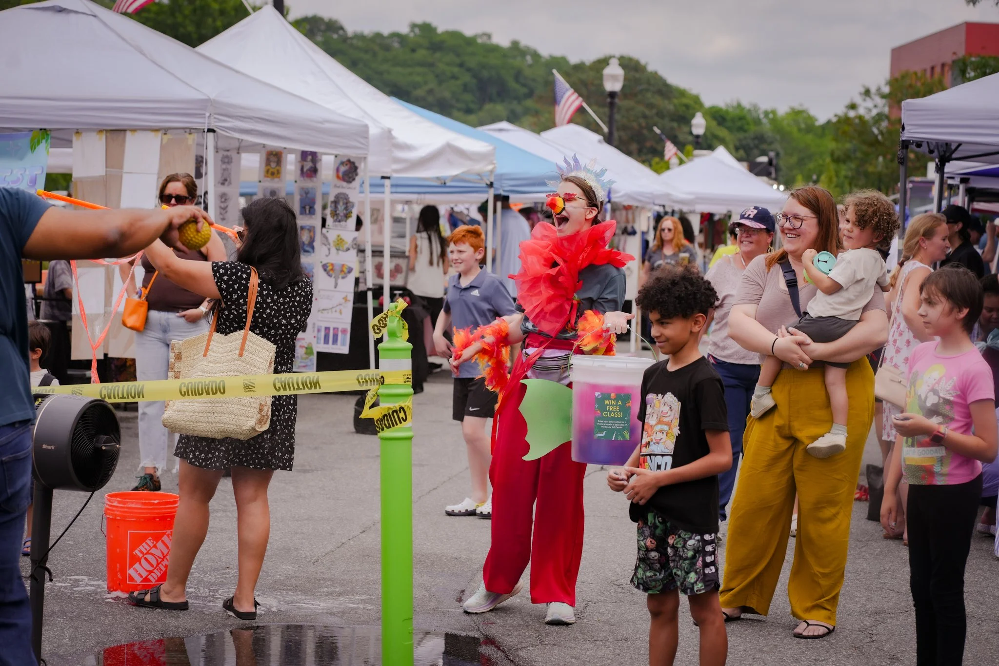 People at an outdoor market or fair, with a clown in red and grey costume, wearing a red nose and a feathered headpiece, entertaining children and adults. Tents and booths line the background, and a caution tape cordons off the area.