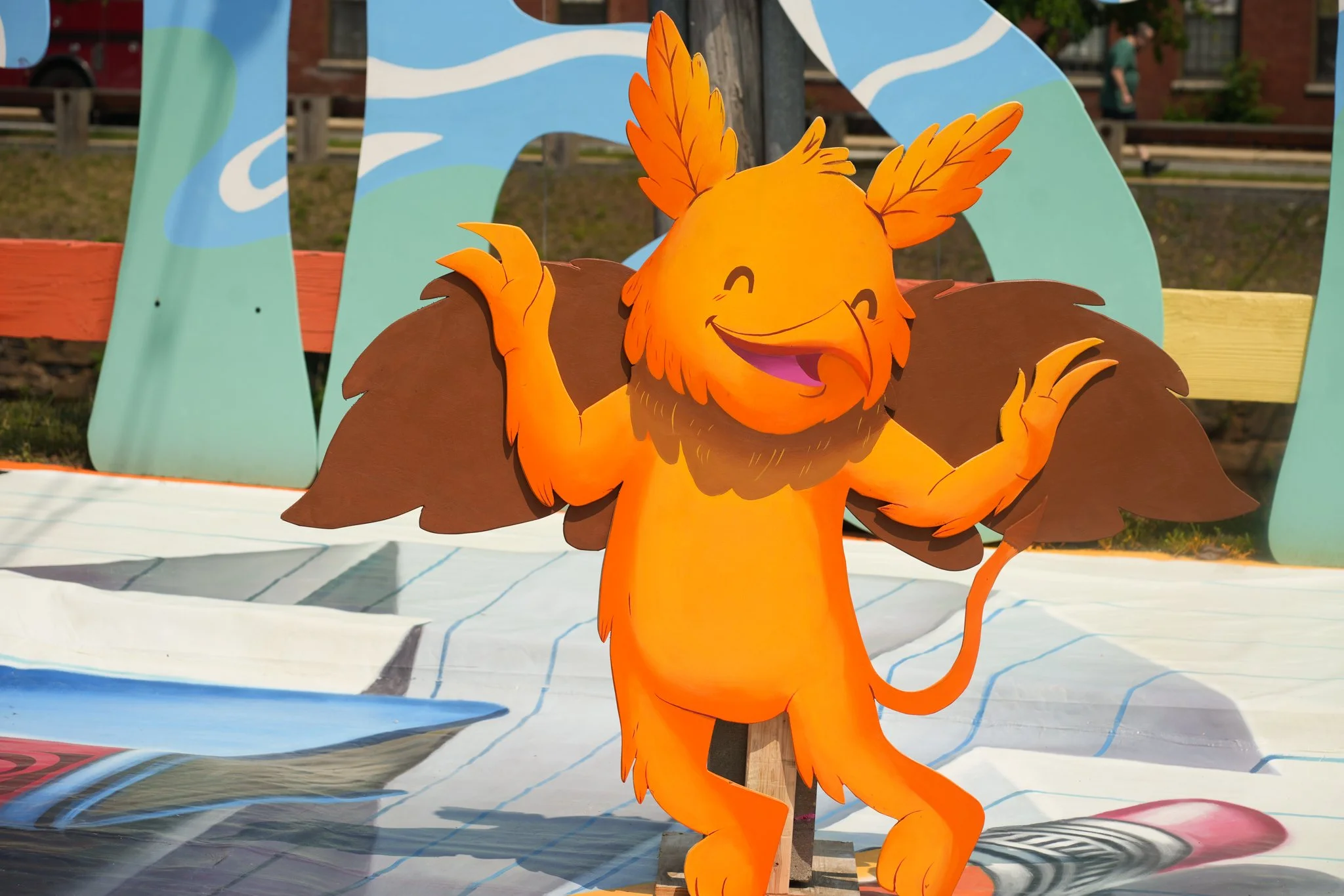 Colorful cartoon lion with a cheerful expression, standing with its arms raised, set against a painted backdrop featuring an outdoor scene.