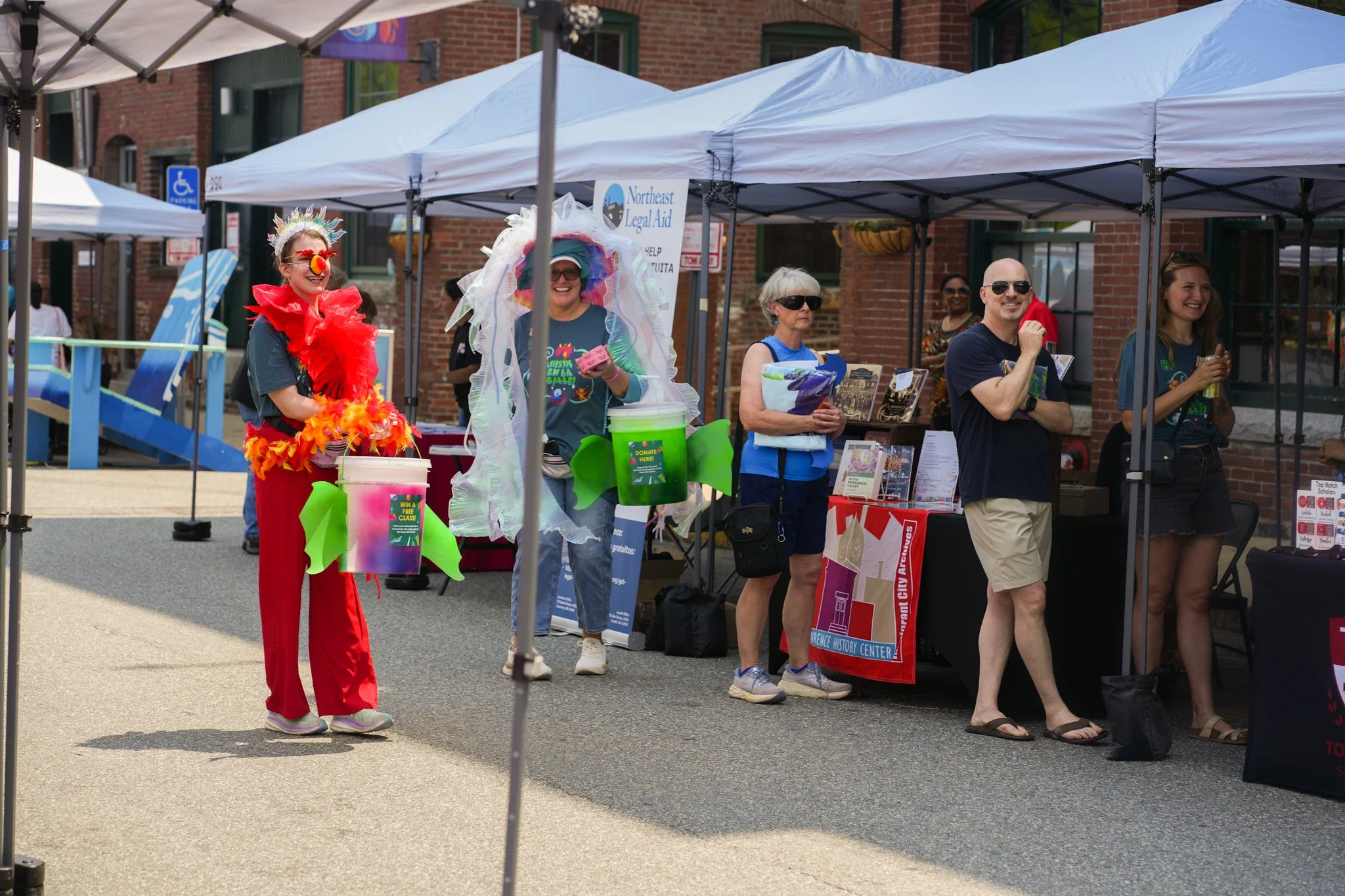 People at an outdoor event booth under canopies, some dressed in colorful costumes, smiling and engaging with each other.