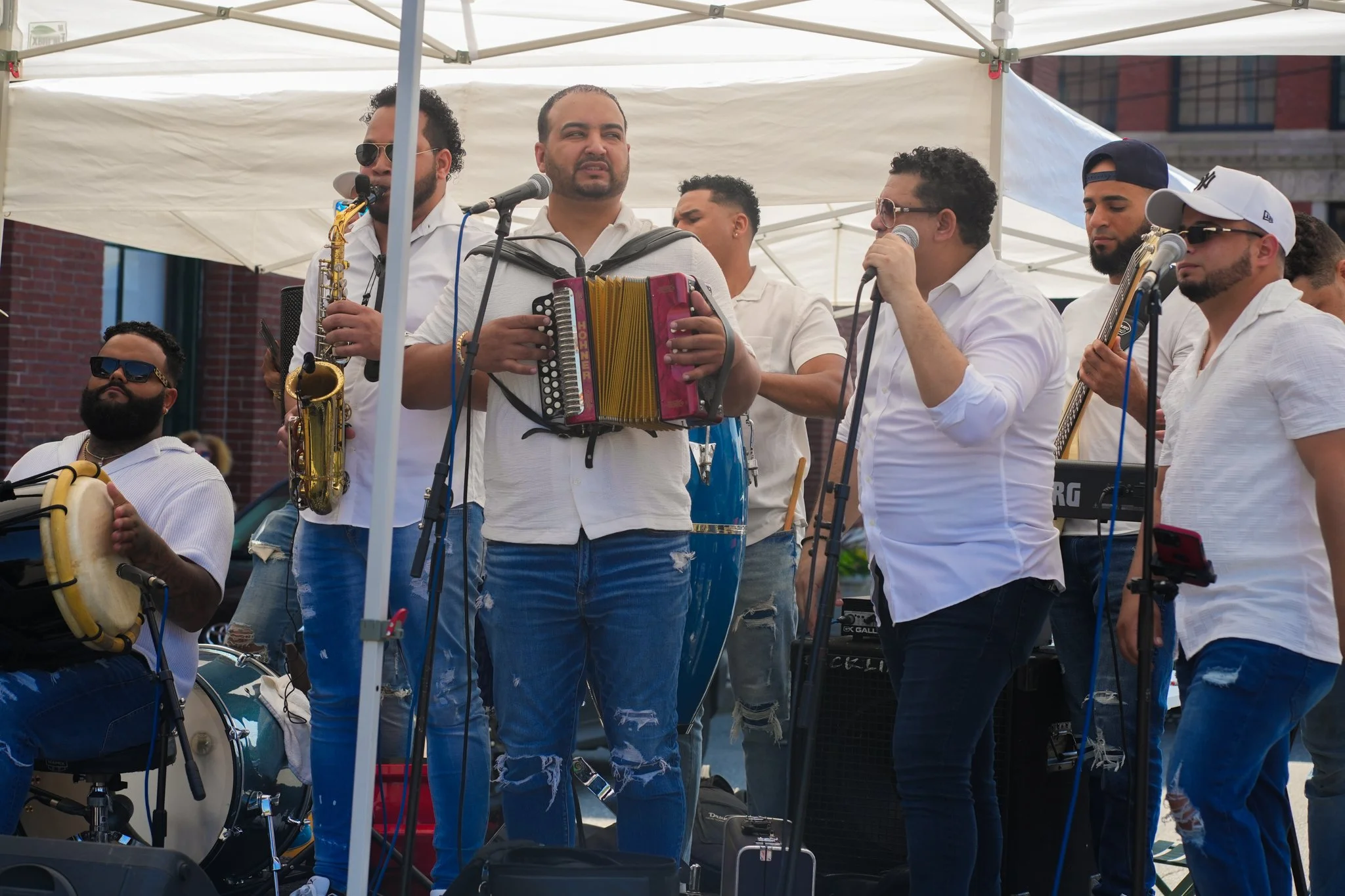 Group of musicians performing outdoors under a white canopy, playing various instruments including a drum, saxophone, accordion, and guitar, dressed in white shirts and jeans.