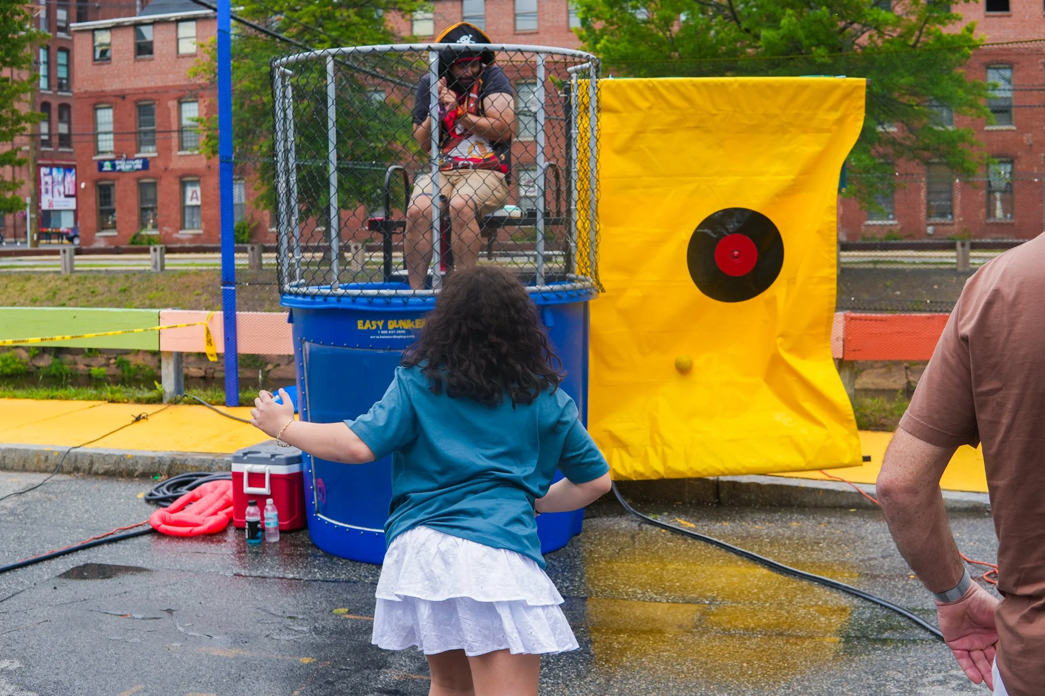 A woman is seated in a dunk tank wearing a pirate hat, while a young girl is throwing a ball at the target to dunk her. There are water and equipment on the ground, and the event takes place outdoors with buildings and trees in the background.