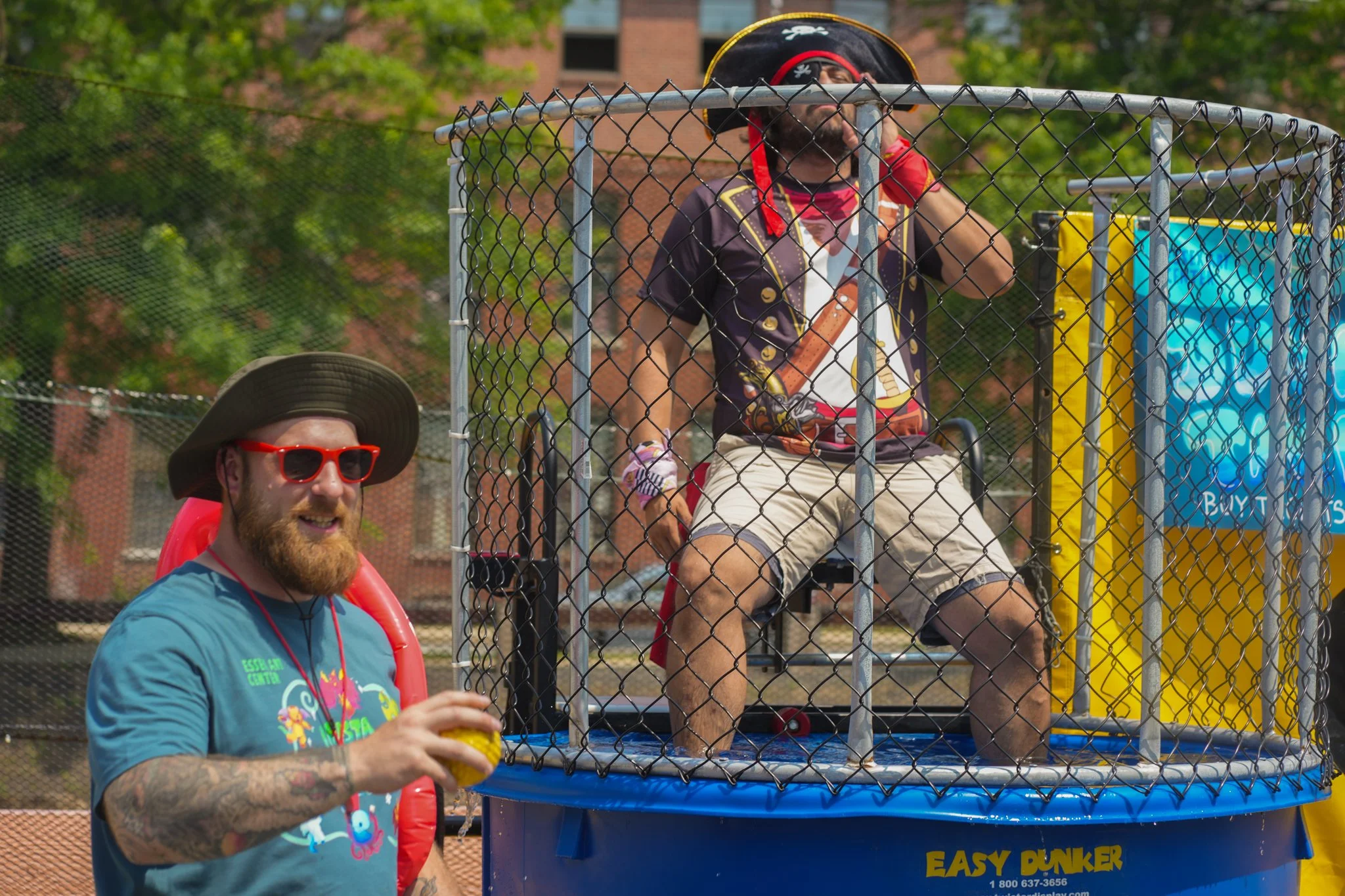 A man dressed as a pirate sitting in a dunk tank, getting soaked, with another man standing outside the tank holding a ball and laughing.