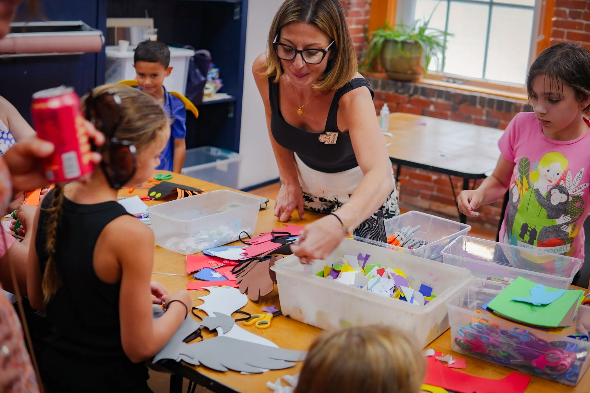 Women and children engaged in arts and crafts activities at a table, with various craft supplies such as paper, scissors, and glue. The setting appears to be a classroom or craft room with brick walls and large windows.