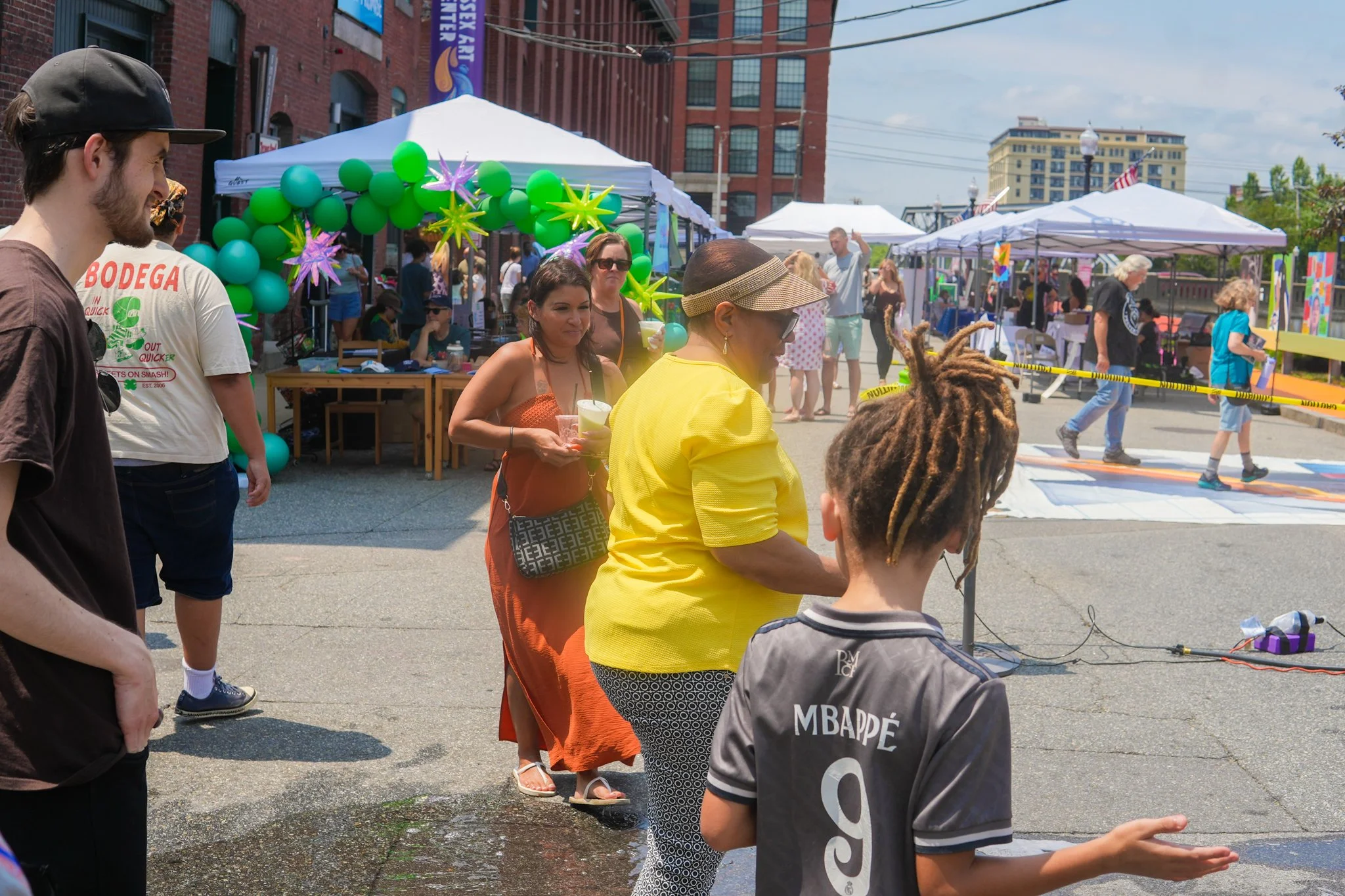 People attending an outdoor festival on a city street, with tents, balloons, and art displays, including children playing on a colorful hopscotch grid.
